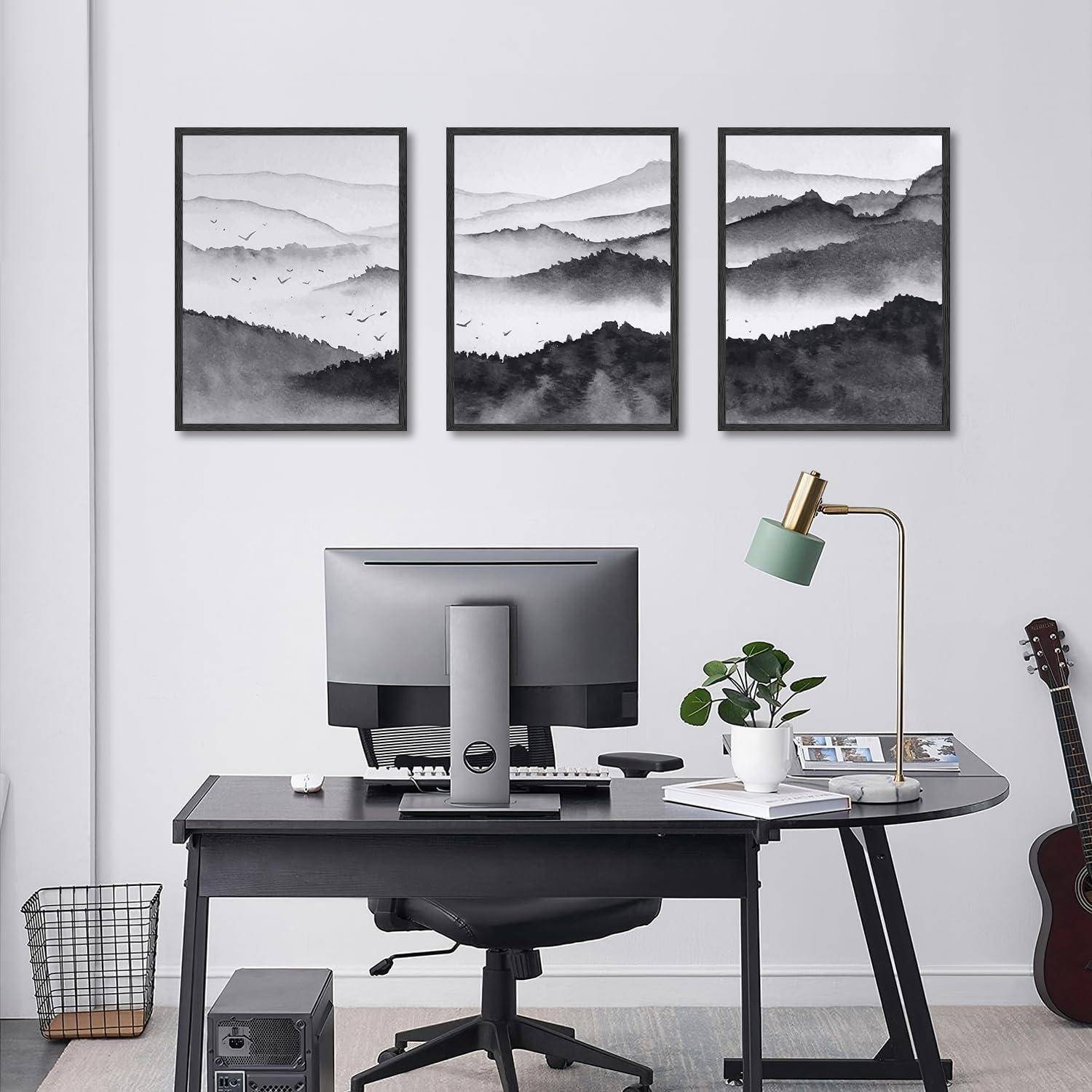 Ein moderner Arbeitsbereich zeigt einen grauen Schreibtisch mit einem Computer, einer Lampe und einer Pflanze. Drei schwarz-weiße Landschaftskunstwerke hängen an der Wand darüber.