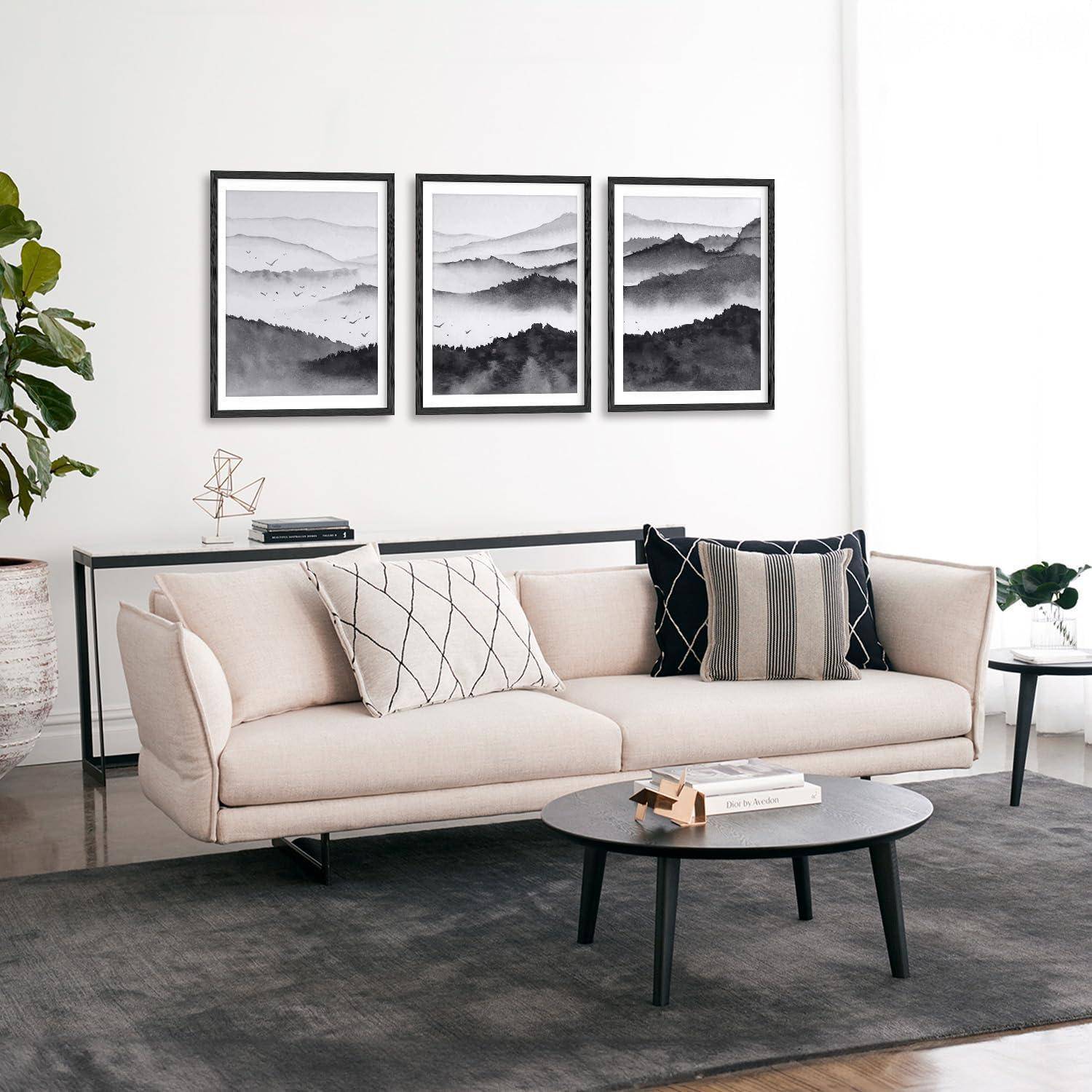 Ein modernes Wohnzimmer verfügt über ein hellbeiges Sofa mit gemusterten Kissen, einen runden Couchtisch und gerahmte schwarz-weiße Bergkunst an der Wand.
