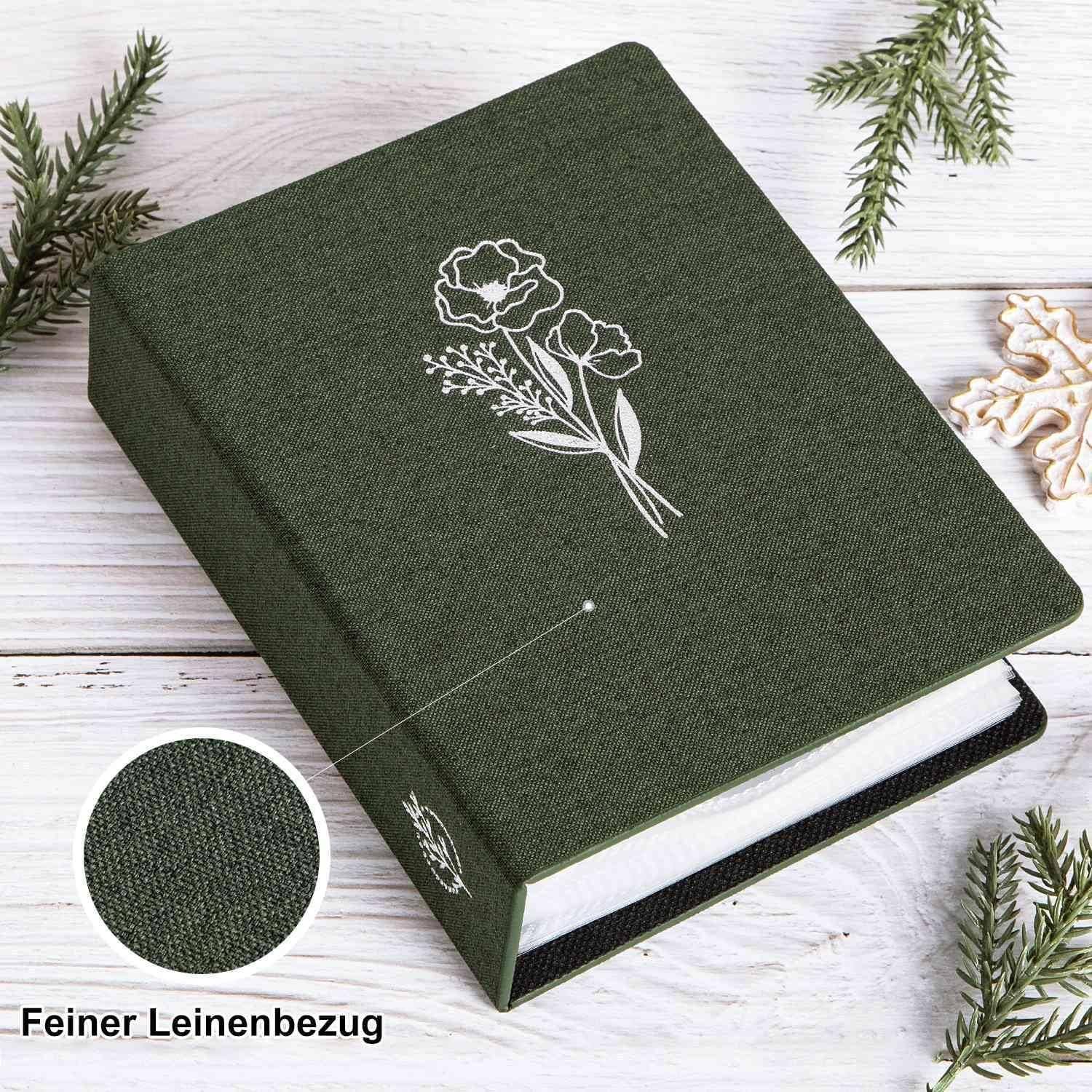 Ein grünes Buch mit Stickerei-Blumenmotiven auf dem Einband, umgeben von Kiefernzweigen auf einer weißen Holzoberfläche. Eine Nahaufnahme zeigt die Leinentextur.