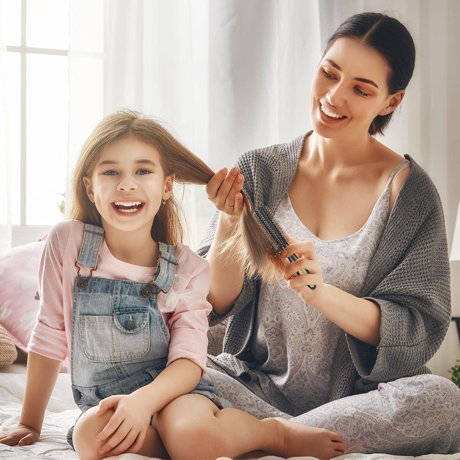 Eine Frau lächelt, während sie einem jungen Mädchen im sonnendurchfluteten Zimmer die Haare bürstet. Das Mädchen, das Jeans-Latzhose trägt, lächelt auch strahlend.