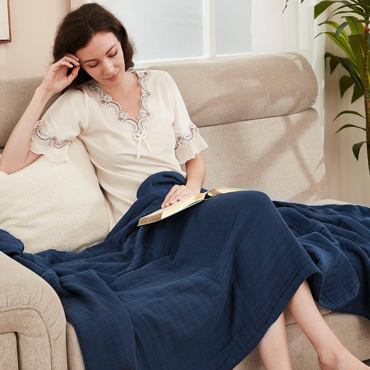 Eine Person entspannt sich auf einem beigefarbenen Sofa und liest ein Buch. Sie ist von einer dunkelblauer Decke bedeckt, und Licht fällt durch ein nahegelegenes Fenster herein.