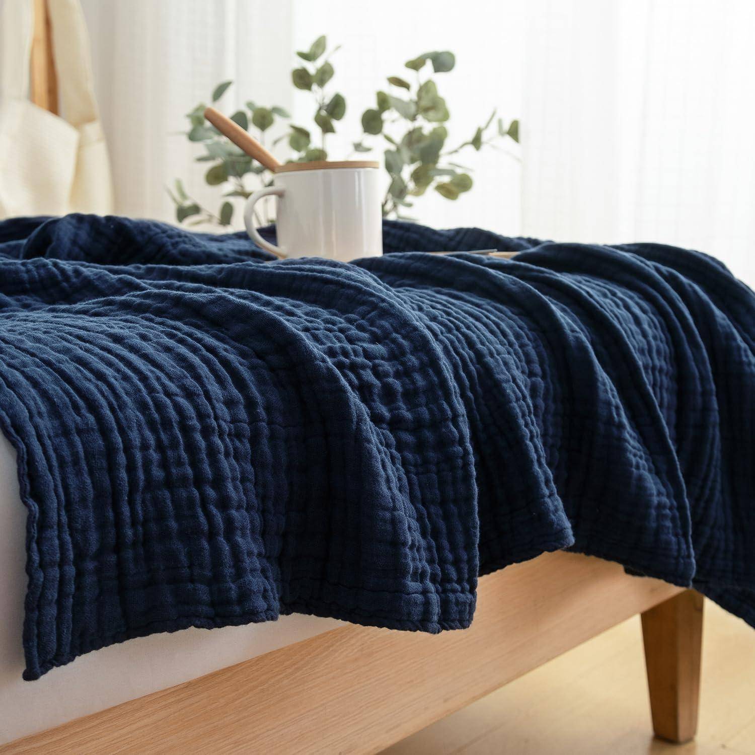 Ein gemütliches Schlafzimmer mit einer blauen strukturierten Decke, die ein Bett bedeckt, einer weißen Tasse und einem Buch oben drauf, und Grünpflanzen im Hintergrund.