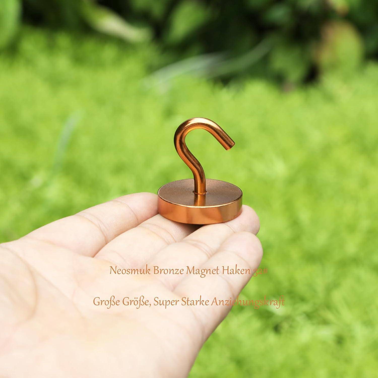 Eine Hand hält einen bronzefarbenen Magnetshaken auf einem grasbewachsenen Hintergrund. Der Haken ist kompakt und metallisch, zum Aufhängen von Gegenständen konzipiert.