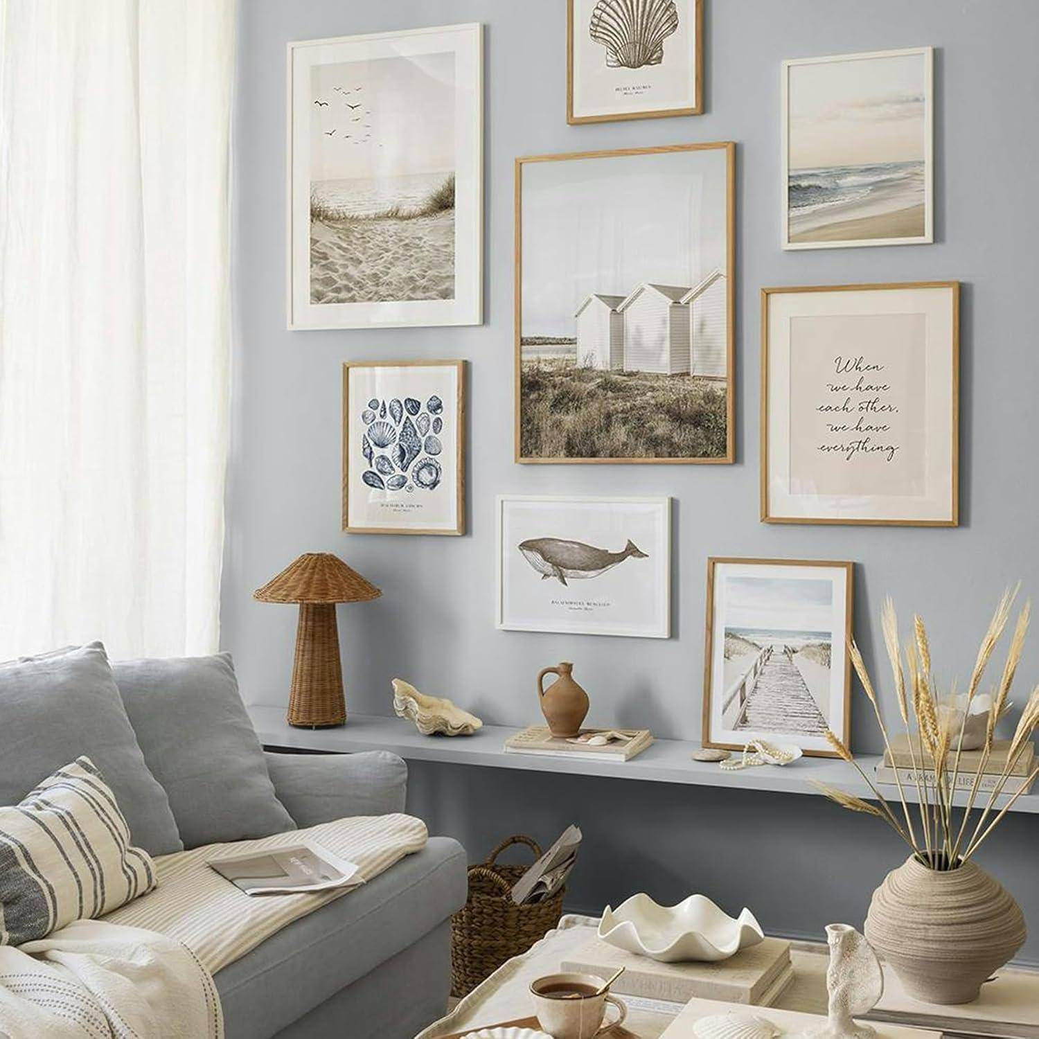 Ein Wohnzimmer mit einer Galerienwand, die Küstenkunst zeigt, ein hellblaugraues Sofa mit Kissen, eine hölzerne Lampe und Dekorationsgegenstände auf Regalen.