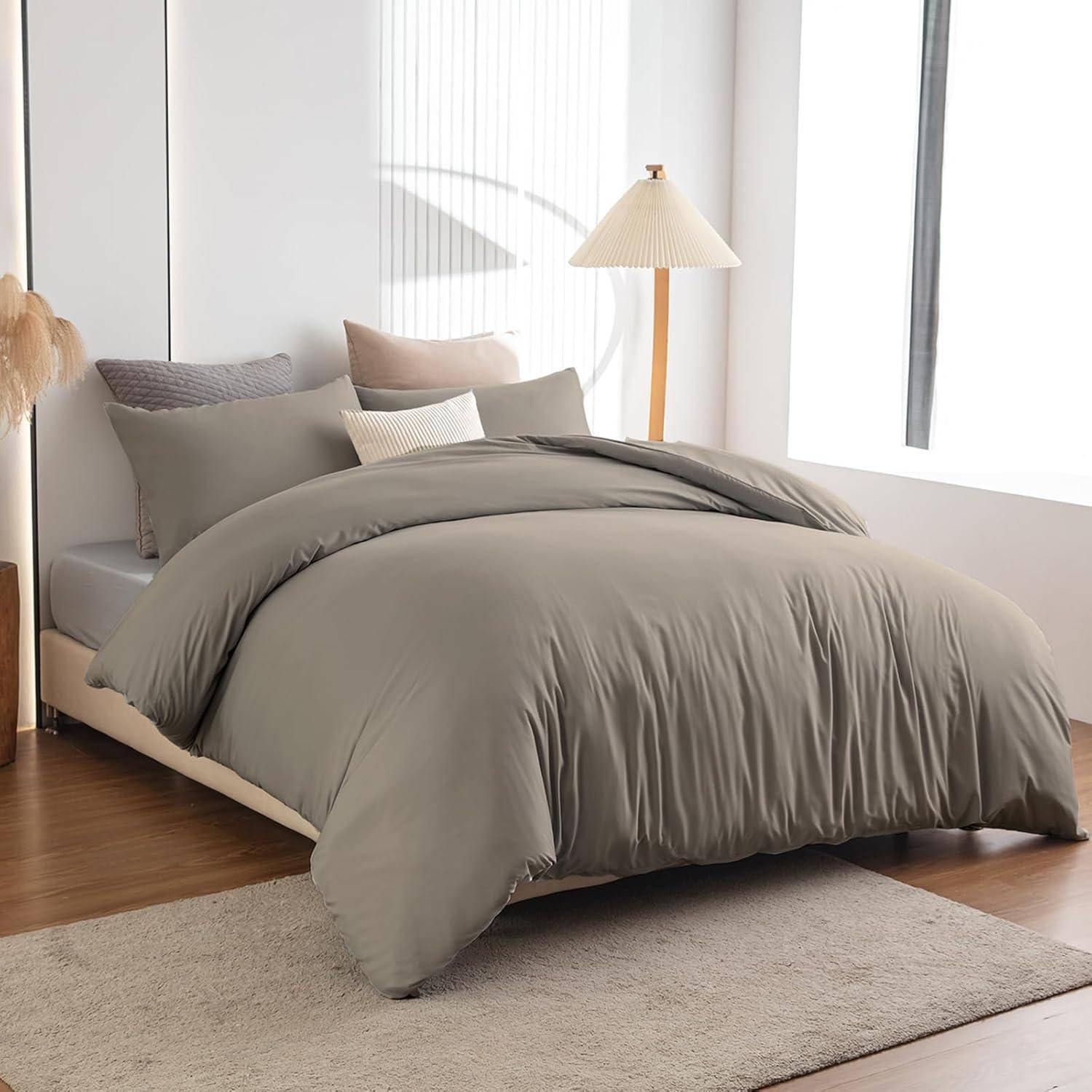 Ein ordentlich gemachtes Bett mit einer weichen grauen Steppdecke, grauen und beigen Kissen und einer modernen Lampe auf einem Holzboden in einem hellen, minimalistischen Zimmer.