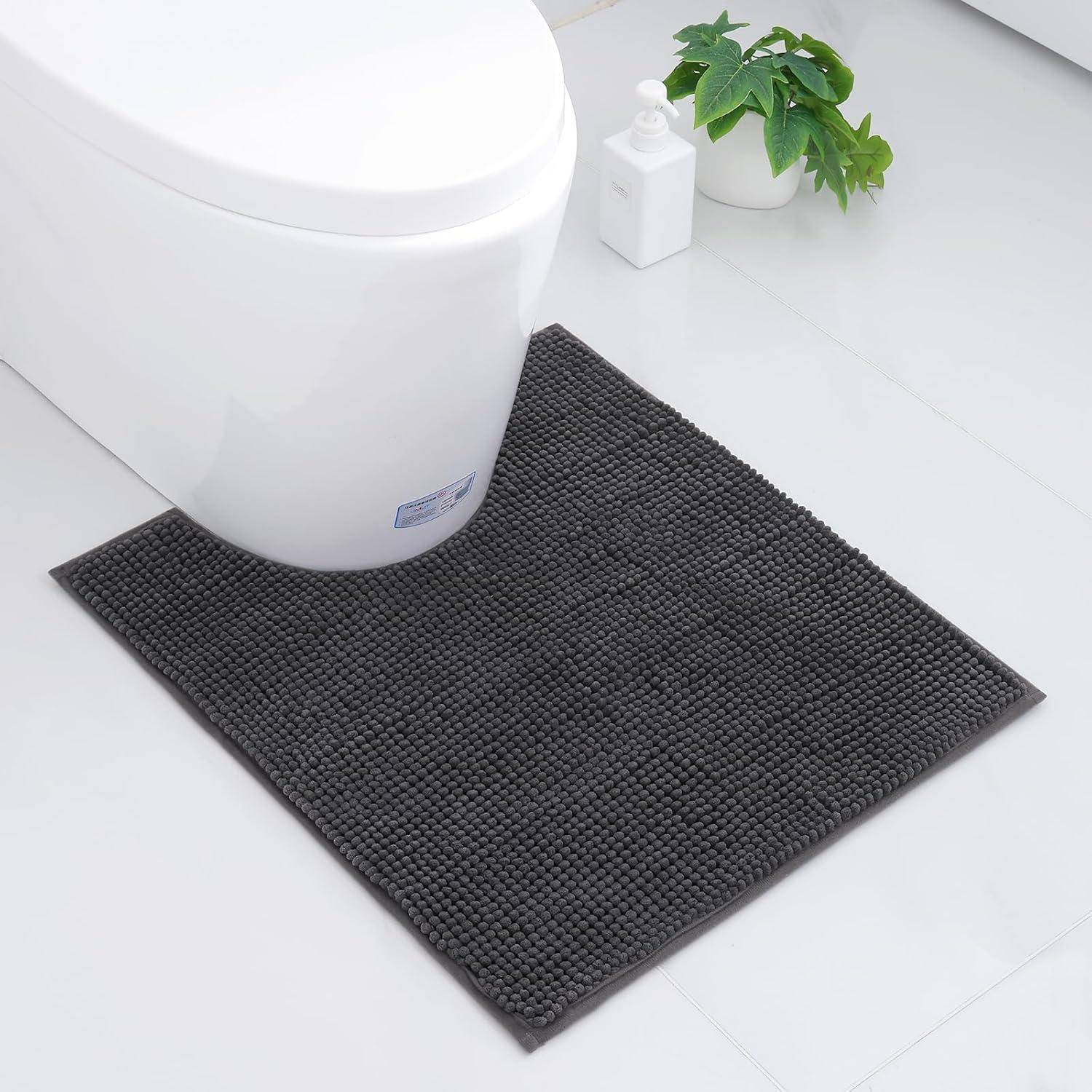 Eine weiße Toilette neben einer dunkelgrauen strukturierten Badmatte. Zwei Pflanzen und ein kleiner Behälter sind auf dem weißen Fliesenboden in der Nähe platziert.