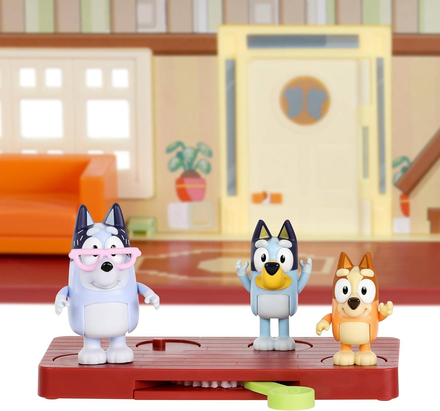 Drei Cartoon-Hundefiguren, eine mit rosa Brille, stehen auf einer Plattform vor einer Spielzeugwohnzimmerszene mit einem Sofa und einer Tür.