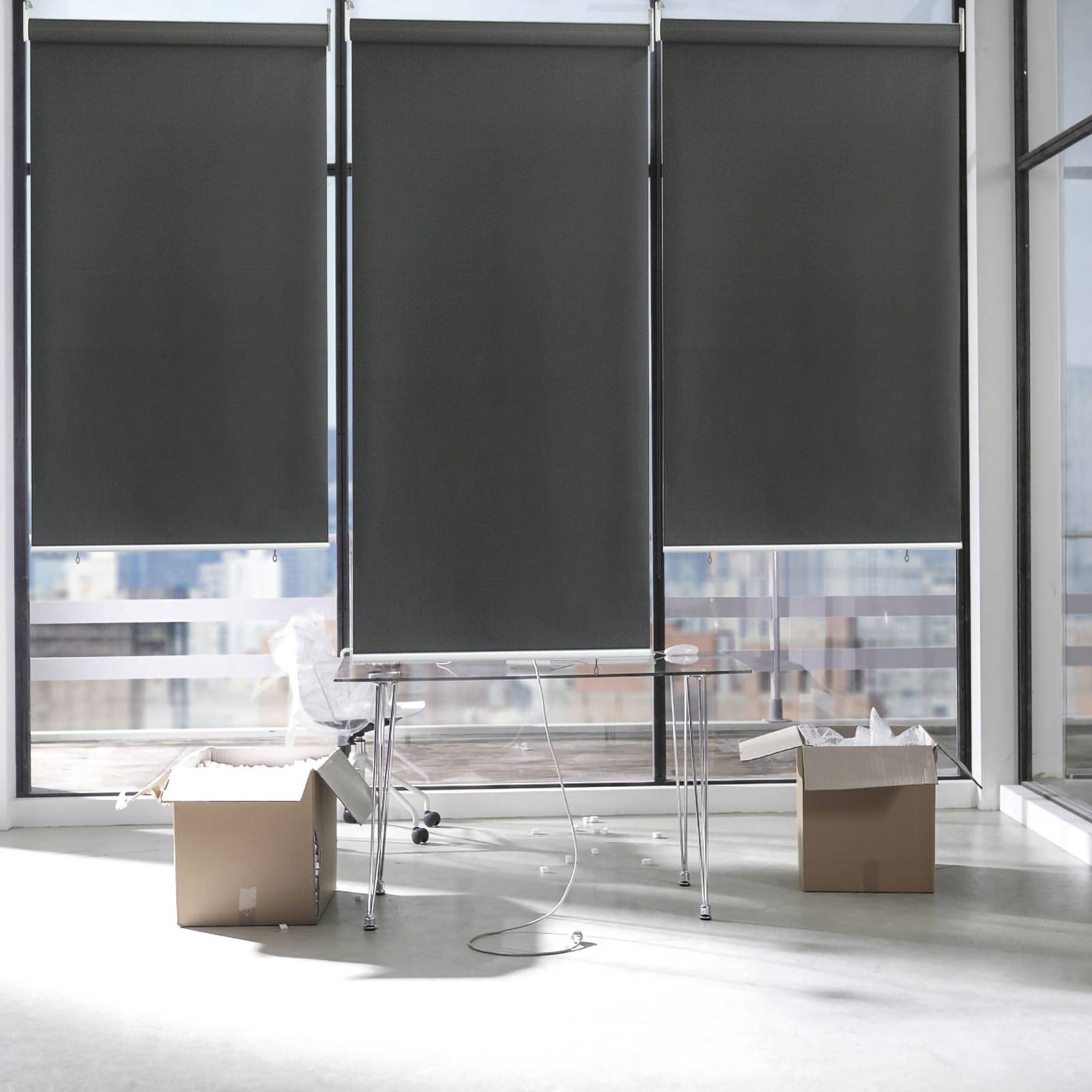 Büroraum mit teilweise herabgelassenen Fensterblenden, einem transparenten Schreibtisch und Kartonboxen auf dem Boden, die auf einen Umzug oder die Einrichtung hindeuten.