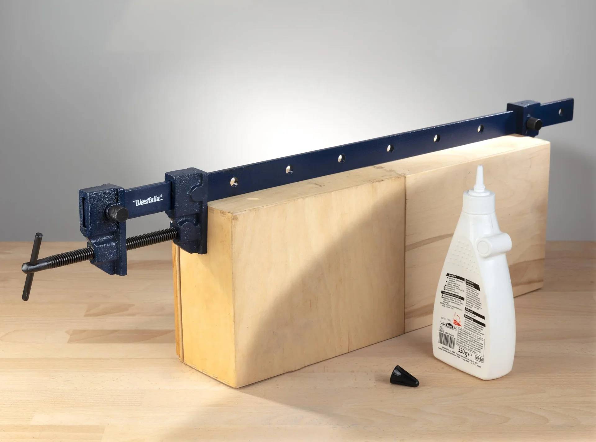 Eine Holzbearbeitungszwinge sichert Holzblöcke auf einem Tisch neben einer Flasche weißem Leim, was auf ein laufendes Tischler- oder DIY-Projekt hindeutet.