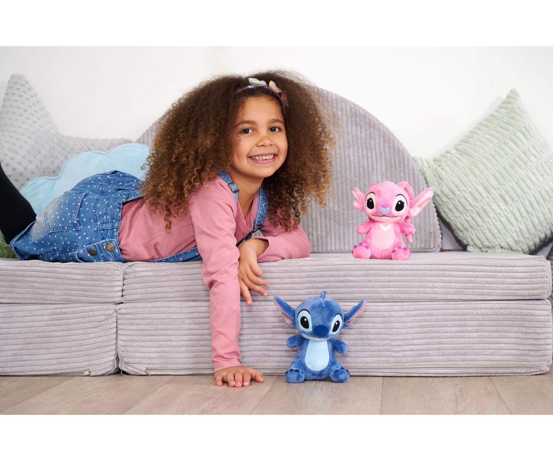 Ein Kind mit lockigem Haar liegt lächelnd auf einem grauen Sofa neben zwei Kuscheltieren, einer rosa und einer blauen Kreatur, und schafft eine verspielte Szene.