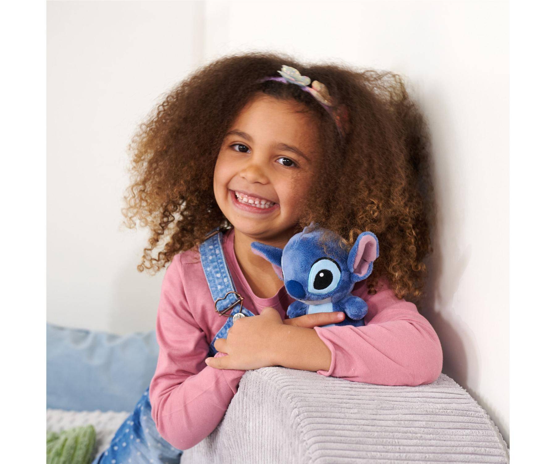 Kind lächelt, während es ein kleines blaues Plüschtier hält. Das Kind hat lockiges Haar und trägt ein rosa Hemd und Jeanoveralls und sitzt auf einem Sofa.