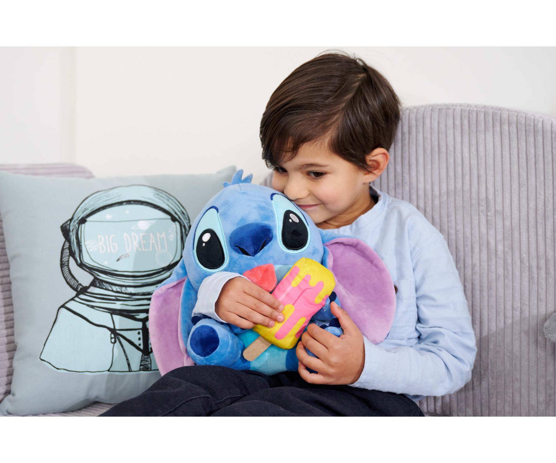 Ein kleines Kind lächelt, während es ein buntes Plüschtier hält und auf einem Sofa sitzt, neben einem Kissen mit einer Astronauten-Illustration.