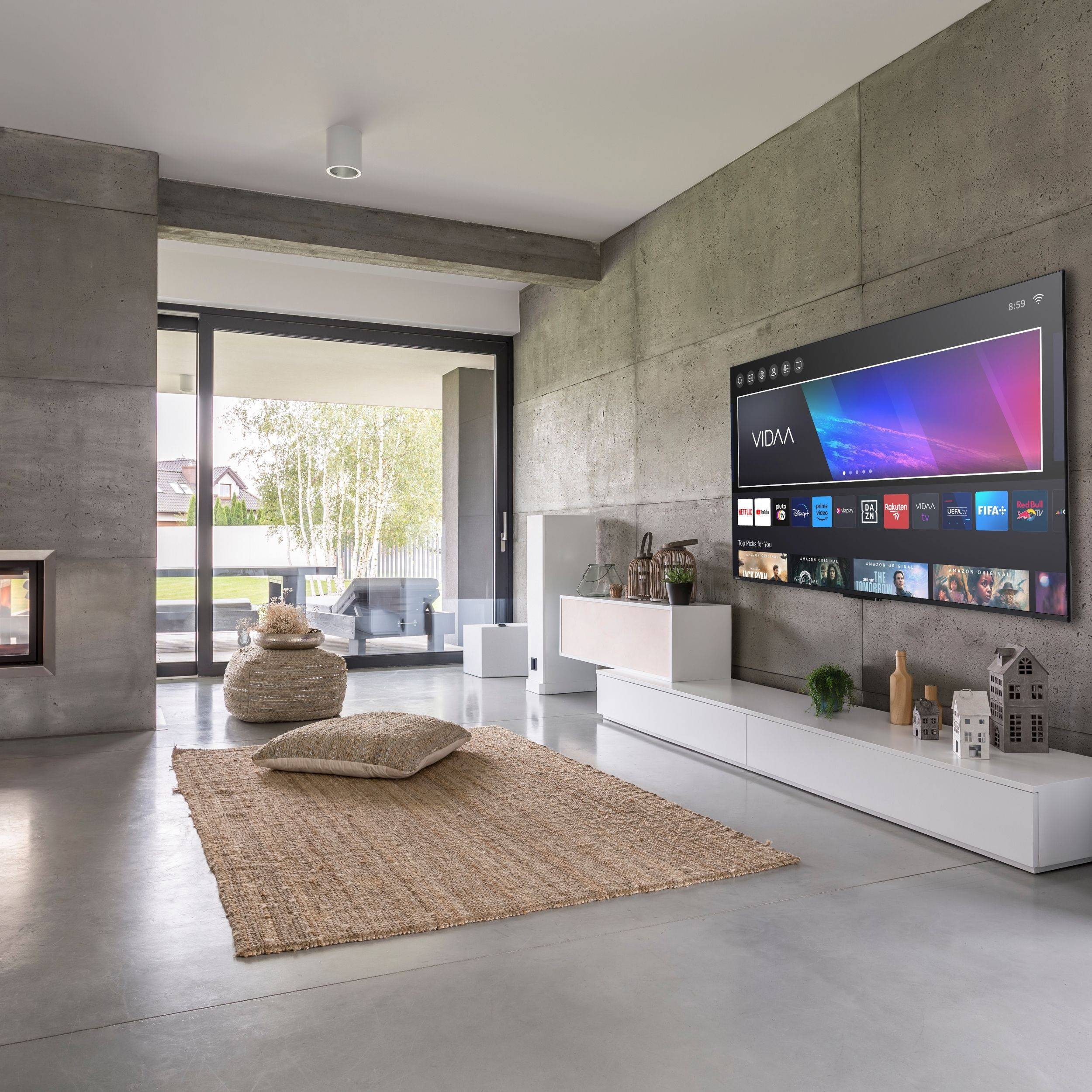 Modernes Wohnzimmer mit Betonwänden, großen Glasschiebetüren und einem an der Wand montierten Smart-TV. Die minimalistische Einrichtung umfasst einen Teppich, ein Kissen und kleine Pflanzen.