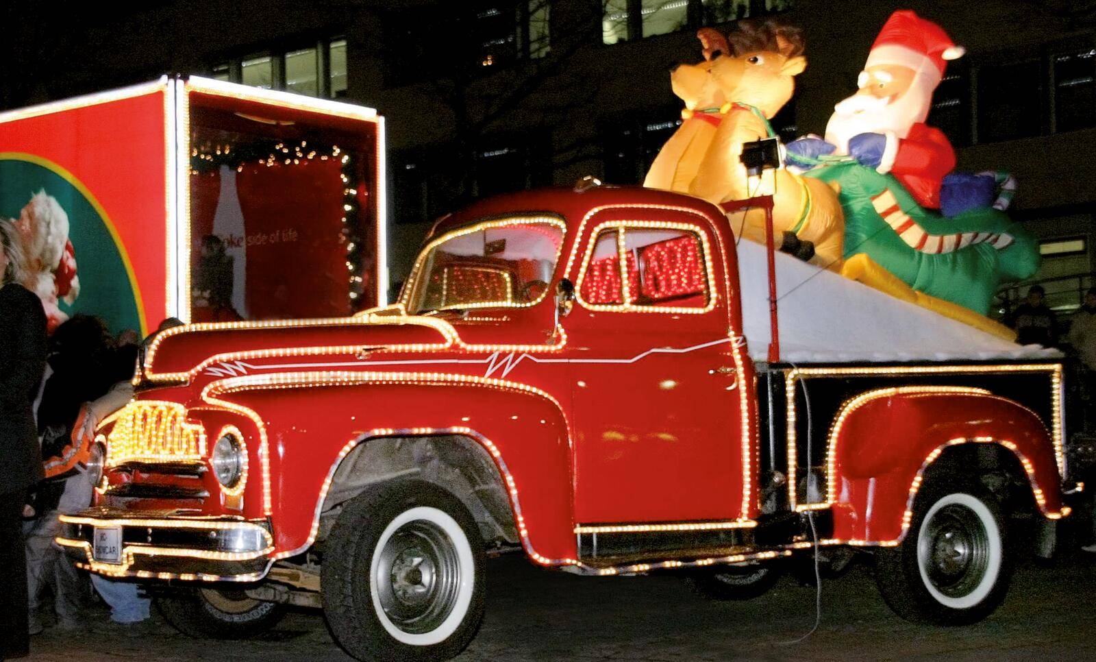 Ein roter Lastwagen, geschmückt mit festlichen Lichtern, transportiert aufblasbare Weihnachtsfiguren, darunter einen großen Weihnachtsmann und zwei Rentiere, während einer Weihnachtsdekoration.