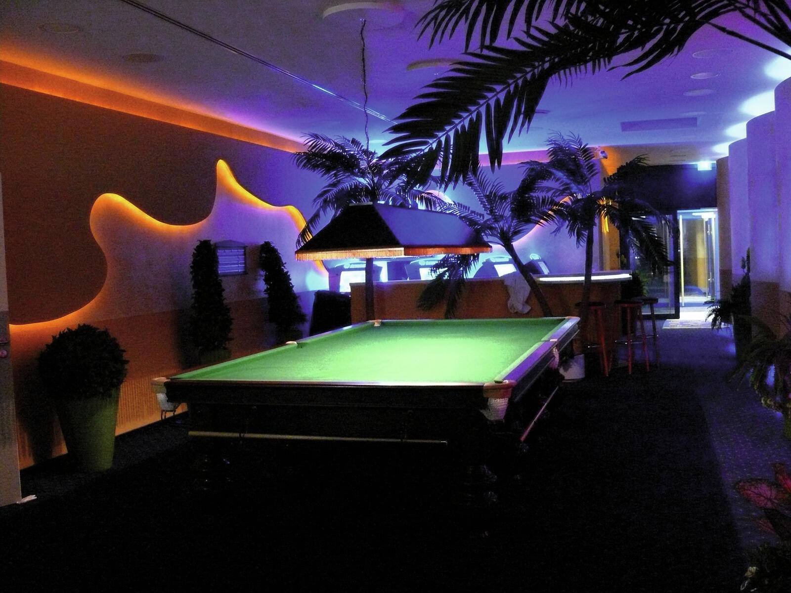 Ein schwach beleuchtetes Billardzimmer mit einem grünen Poolbillardtisch, tropischen Pflanzen und warmem Licht schafft eine entspannende Atmosphäre.