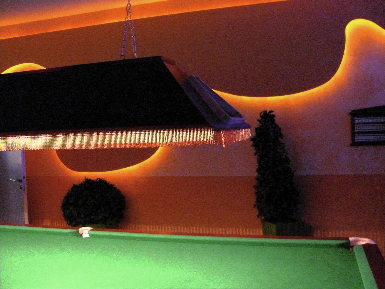 Ein schwach beleuchtetes Billardzimmer mit einem grünen Pooltisch, einem Kronleuchter mit Fransen an der Decke und dekorativen Pflanzen vor einer orange beleuchteten Wand.