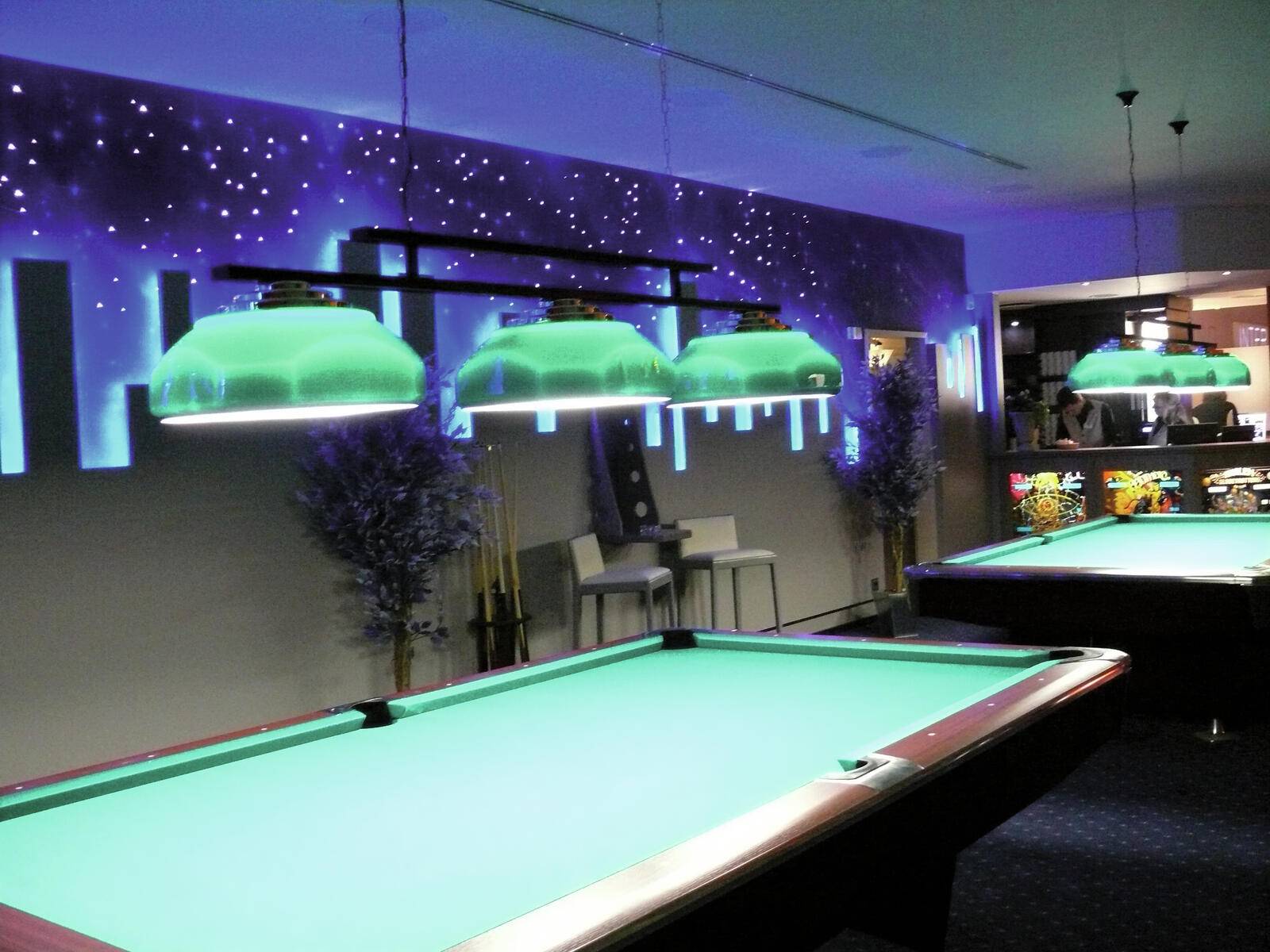 Ein schwach beleuchtetes Billardzimmer mit beleuchteten Billardtischen, moderner Einrichtung, einem Sternendesign an der Wand und einem Sitzbereich im Hintergrund.