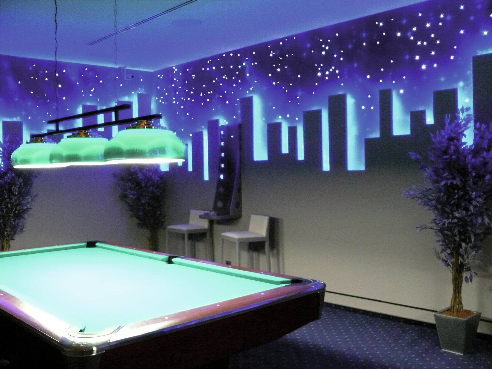 Ein Billardtisch steht in einem schwach beleuchteten Raum. An den Wänden wird eine leuchtende Stadtlandschaft mit Sternen angezeigt, die eine nächtliche Atmosphäre schafft. Zwei Stühle stehen in der Nähe.