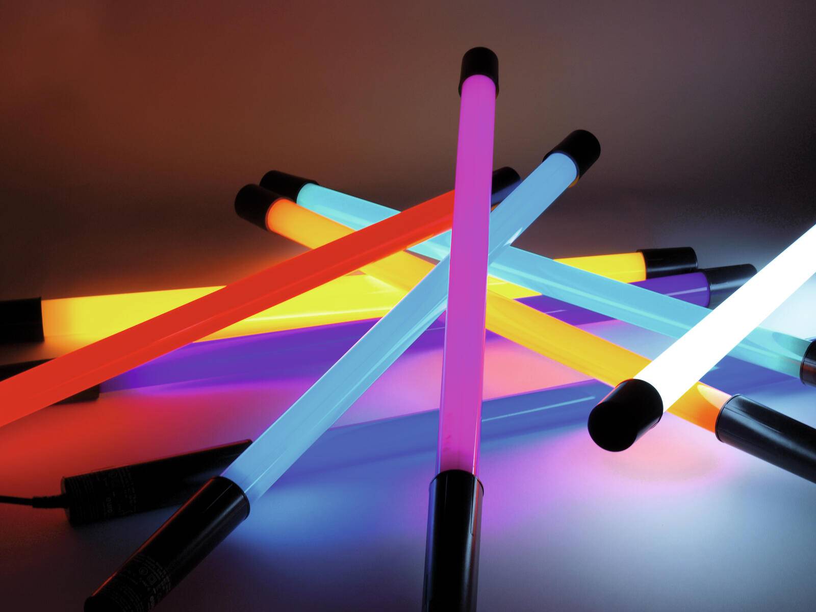 Farbenfrohe Leuchtstäbe, die blaue, rosa, orange und gelbe Töne ausstrahlen, sind über einer reflektierenden Oberfläche angeordnet und erzeugen einen lebendigen visuellen Effekt.