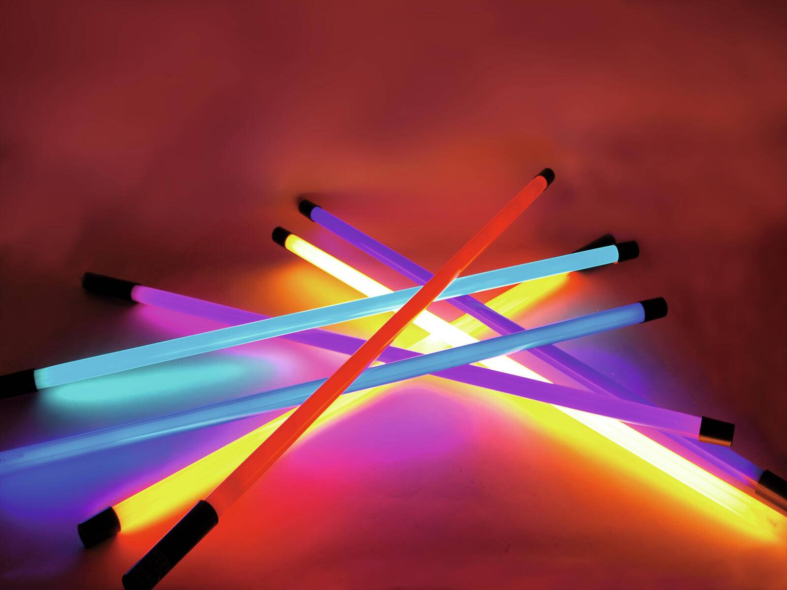 Mehrere leuchtende Neonlichtstäbe in Pink, Blau und Orange, die sich vor einem dunklen Hintergrund überschneiden und ein lebendiges, farbenfrohes Muster erzeugen.