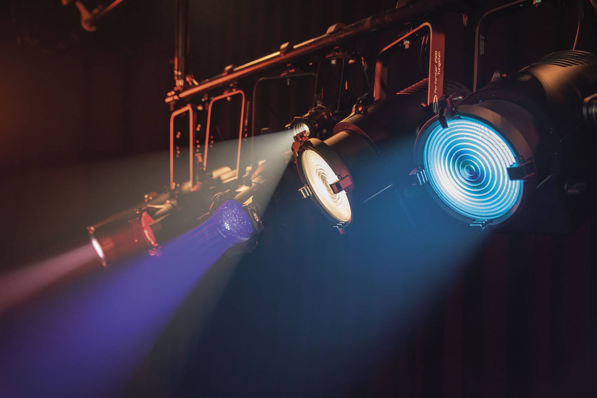 Eine Reihe von Bühnenscheinwerfern, mit einem markanten blauen Scheinwerfer, erleuchtet einen dunklen Aufführungsraum und schafft eine konzentrierte, dramatische Atmosphäre.