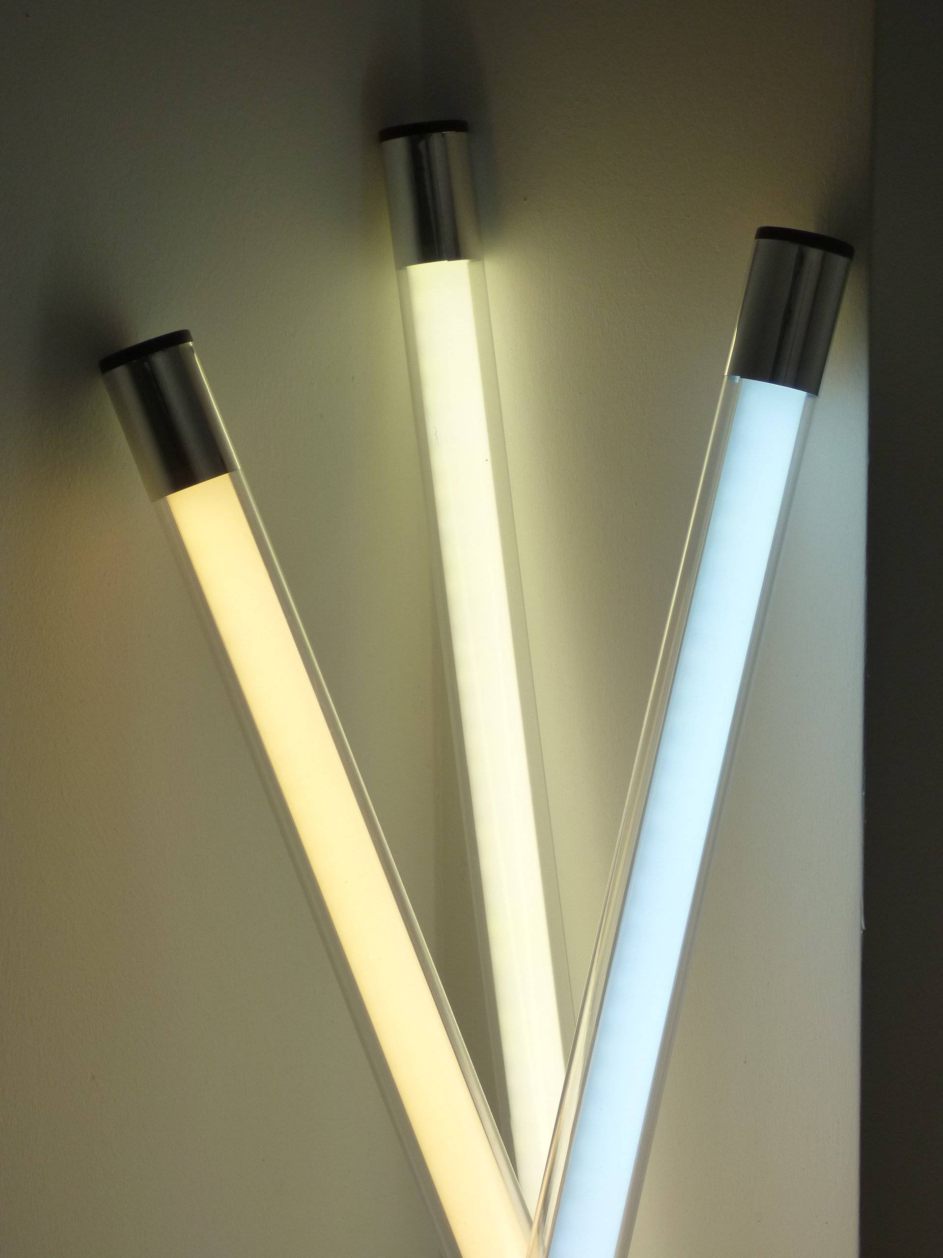Drei vertikale LED-Leuchtstoffröhren mit metallischen Kappen, die an einer Wand montiert sind, wobei jede eine andere Farbe ausstrahlt: Warmweiß, Neutralweiß und Kühlblau.