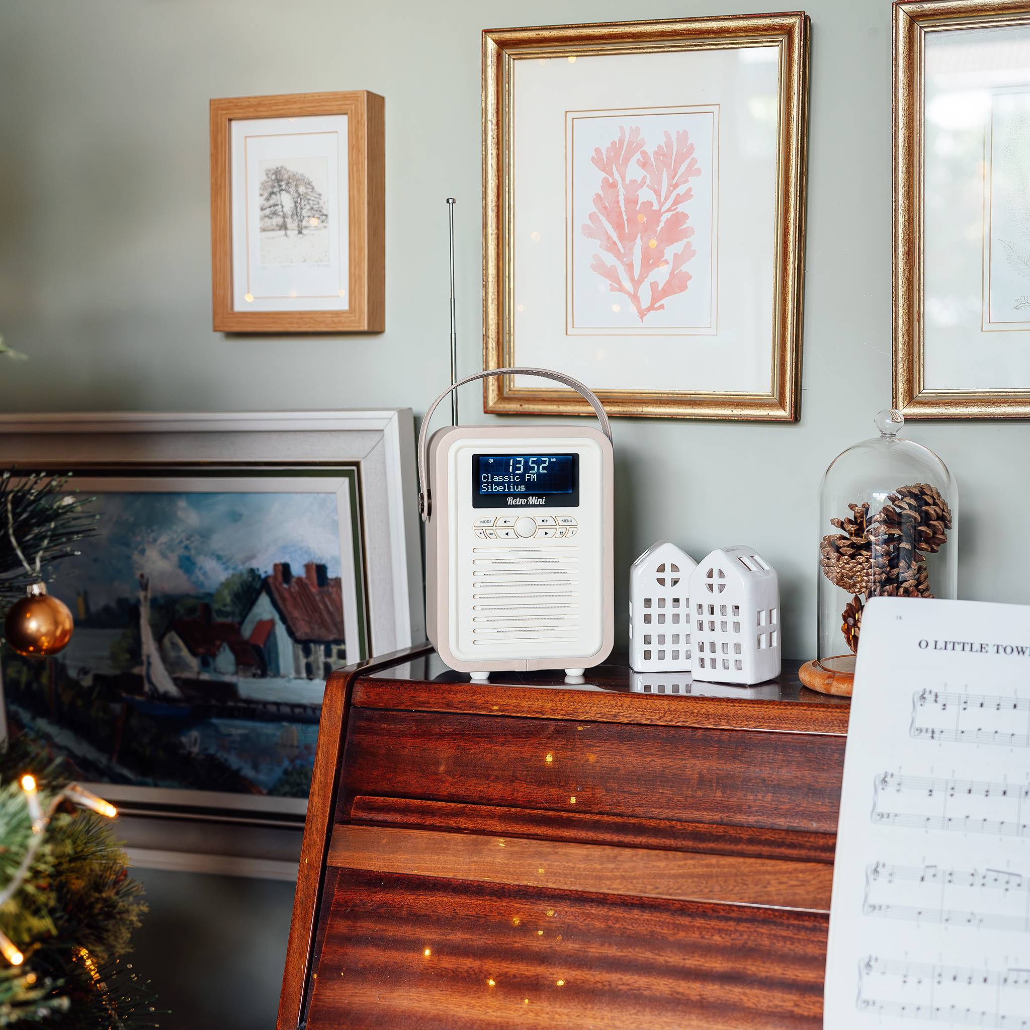 Ein Radio sitzt auf einem Holzregal, umgeben von Bilderrahmen mit Kunstwerken und dekorativen Gegenständen, einschließlich eines kleinen Modellhauses und getrockneter Blumen.