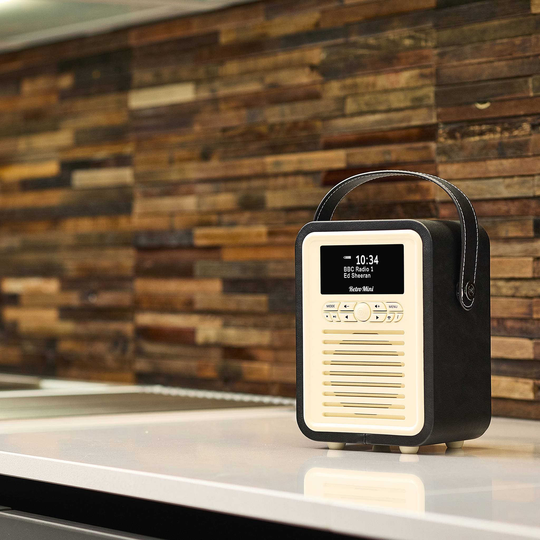 Ein Radio im Retro-Stil mit digitaler Anzeige, die '10:34' und 'BBC Radio 1' zeigt. Es ruht auf einer modernen Küchenarbeitsplatte mit einer Holzrückwand.