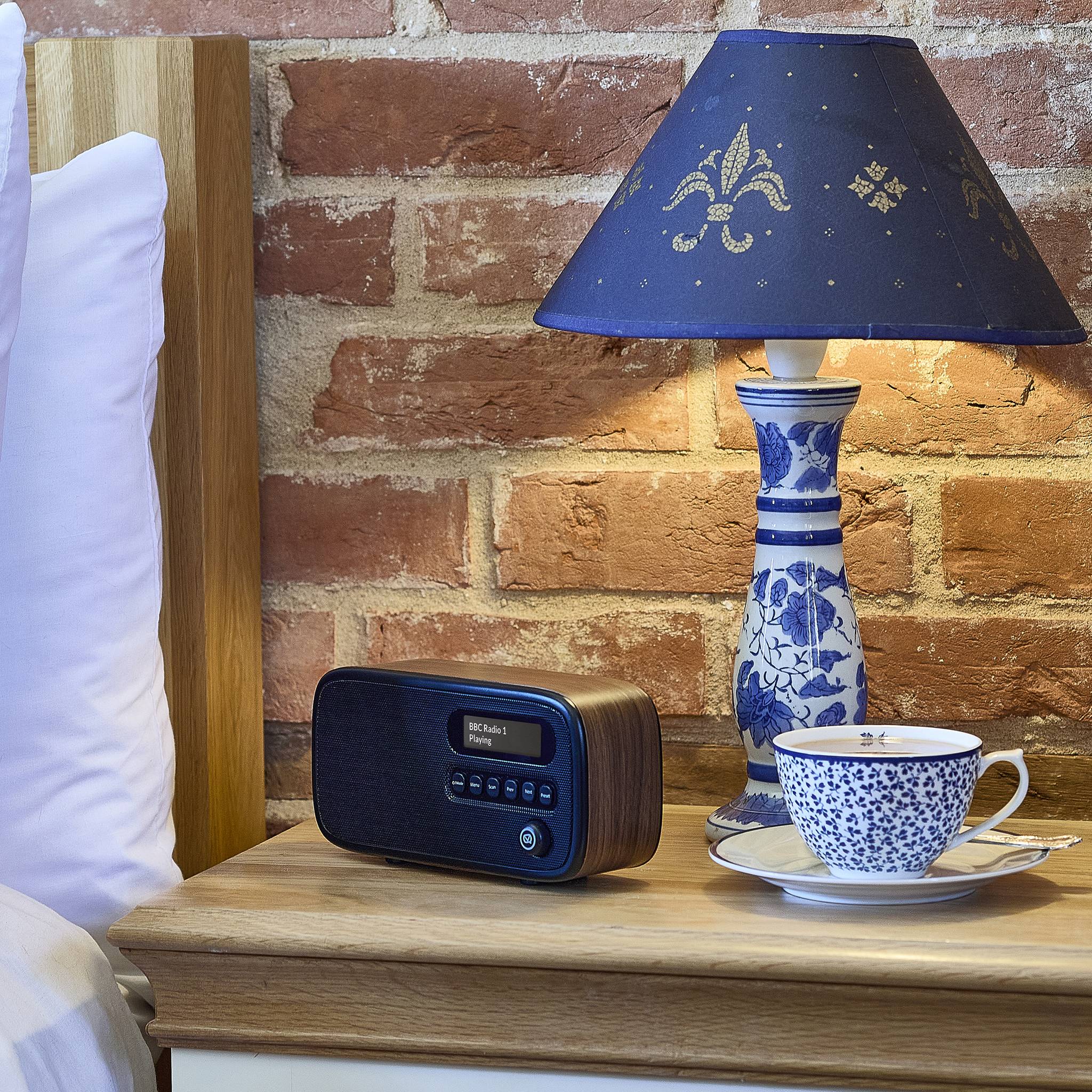 Ein Nachttisch mit einer verzierte Lampe, einem digitalen Uhrenradio und einer blaugemusterten Teetasse auf einer Untertasse vor einem Backsteinmauer-Hintergrund.