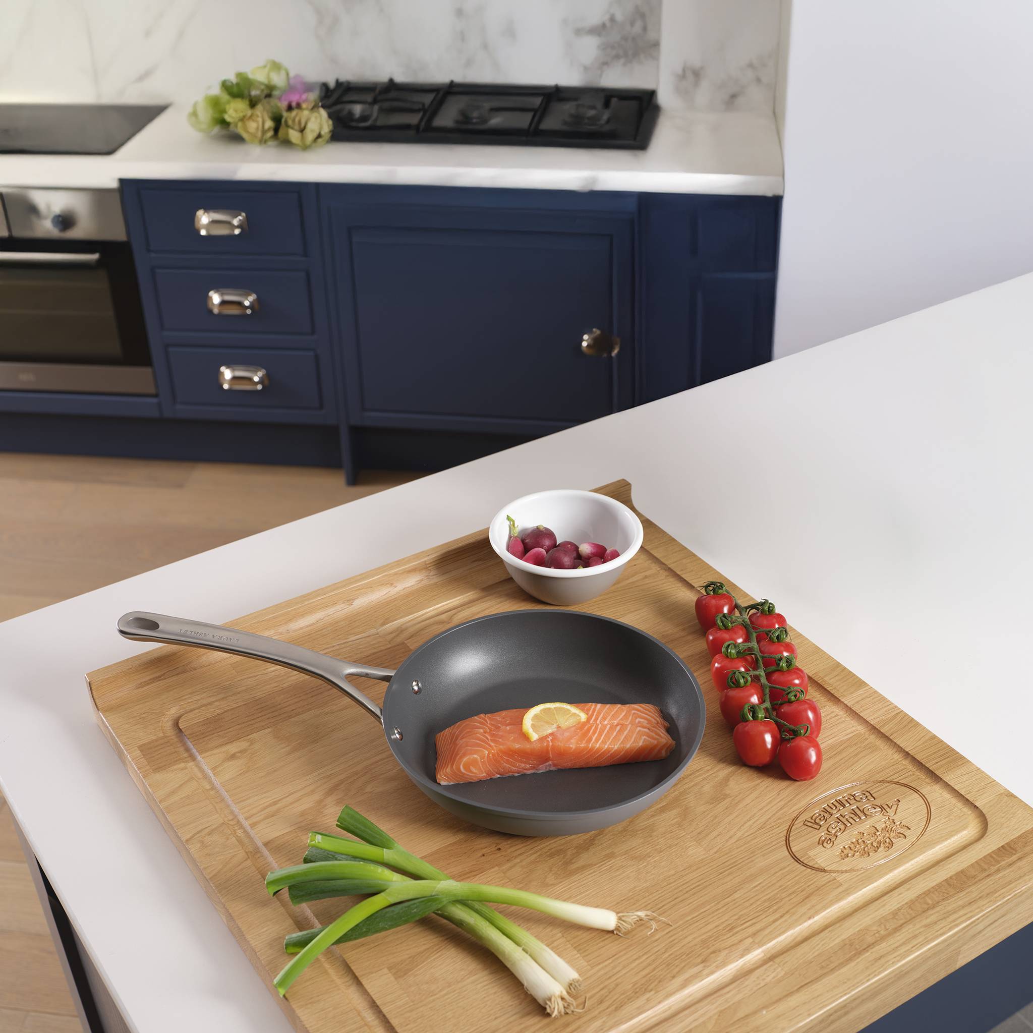 Ein Holzschneidebrett auf einer Küchentheke hält eine Pfanne mit Lachs, Kirschtomaten, Frühlingszwiebeln und Radieschen, mit einem Herd in der Nähe.