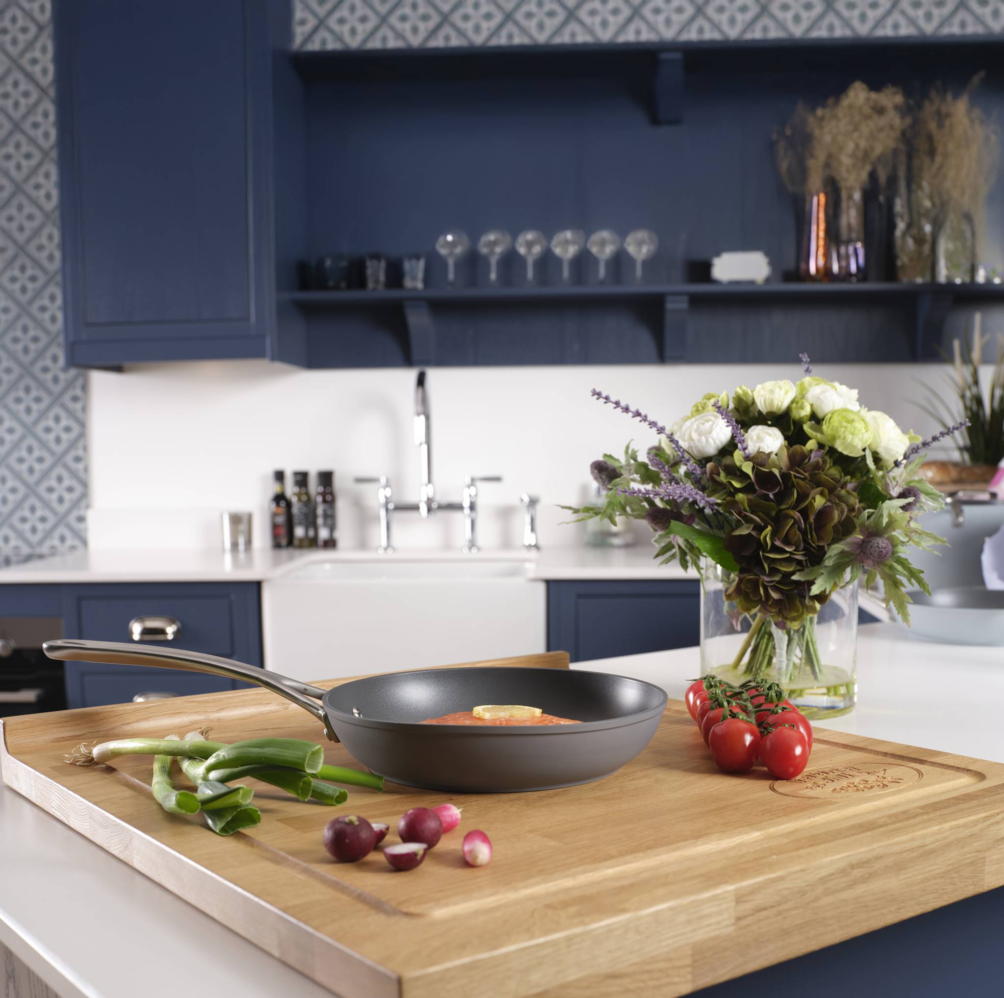 Eine moderne Küche mit blauen Schränken zeigt ein Schneidebrett mit Zwiebeln und eine Bratpfanne. Eine Blumenvase verziert die Arbeitsfläche.