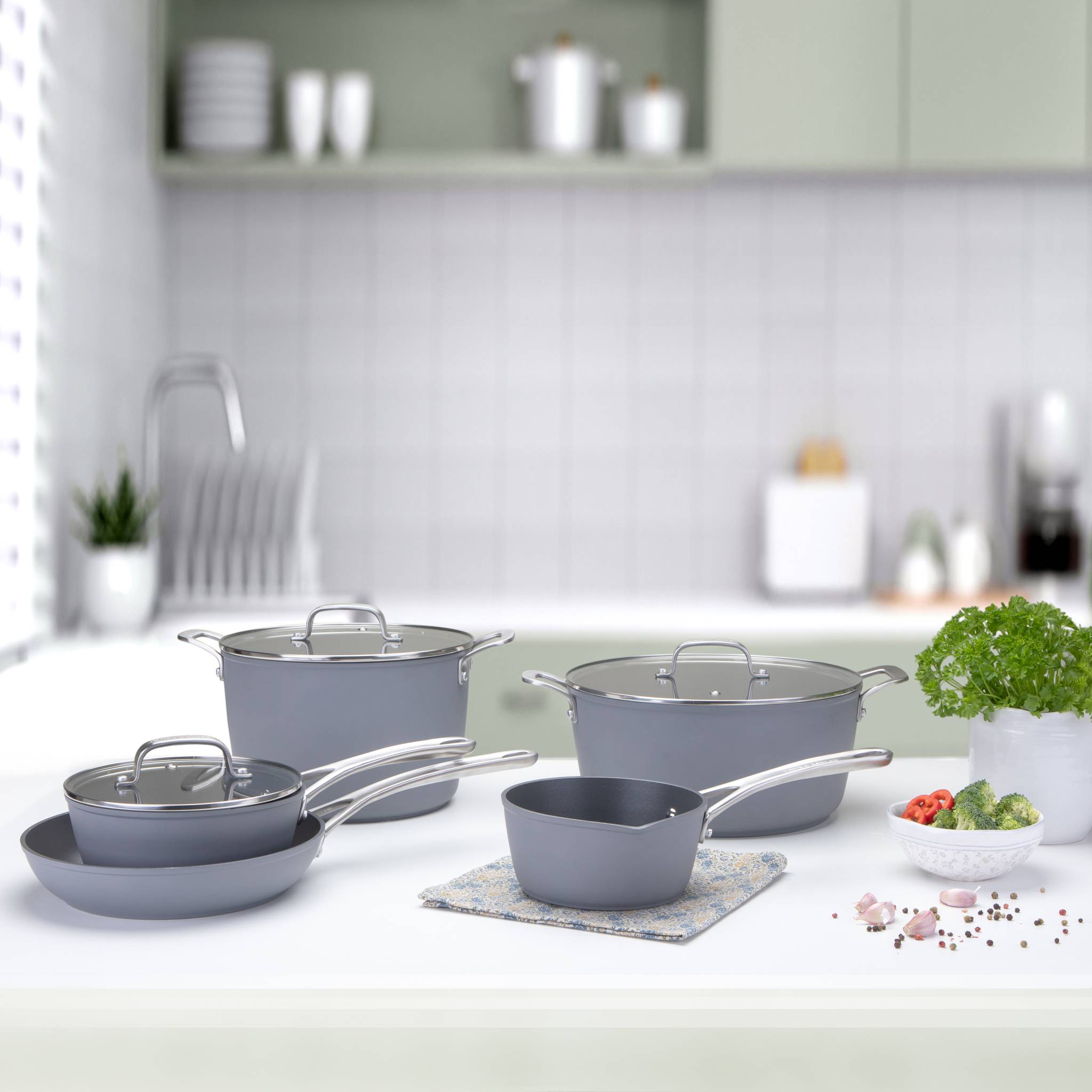 Ein Set aus grauem Kochgeschirr, bestehend aus Töpfen und Pfannen mit Glasdeckeln, auf einer Küchenarbeitsplatte angeordnet, mit einer Topfpflanze in der Nähe.