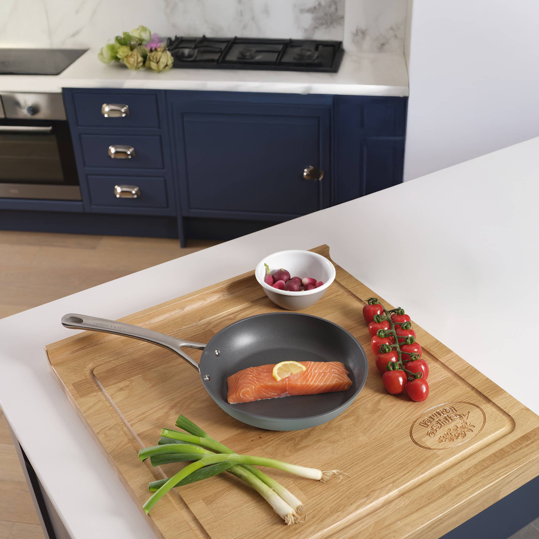 Eine Küchenszene, die ein Lachsfilet in einer Bratpfanne auf einem Holzbrett zeigt, umgeben von Zwiebeln, Radieschen und Kirschtomaten.