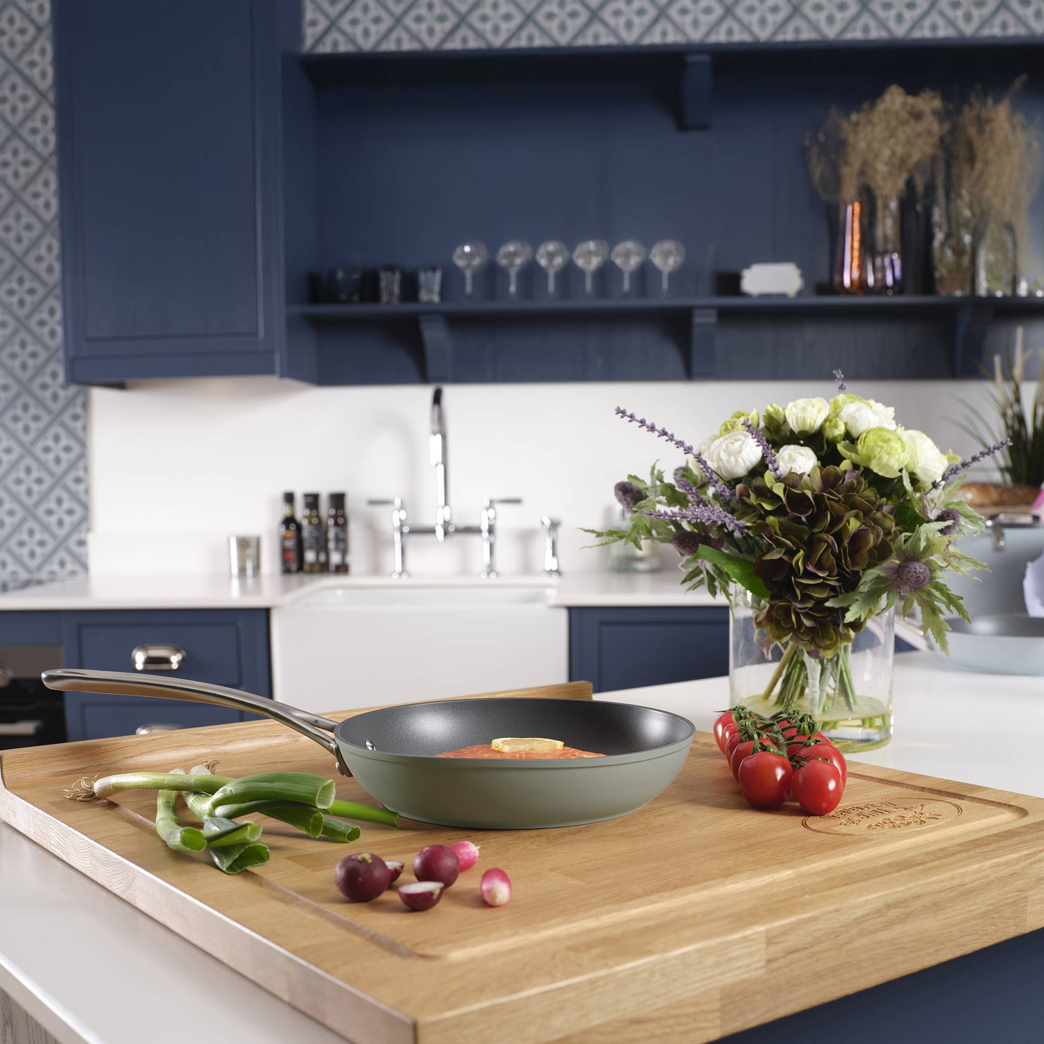 Eine Küche mit blauen Schränken und einer Holzarbeitsplatte zeigt eine Pfanne mit Eiern, grünen Zwiebeln und Radieschen, neben einer Blumenvase.