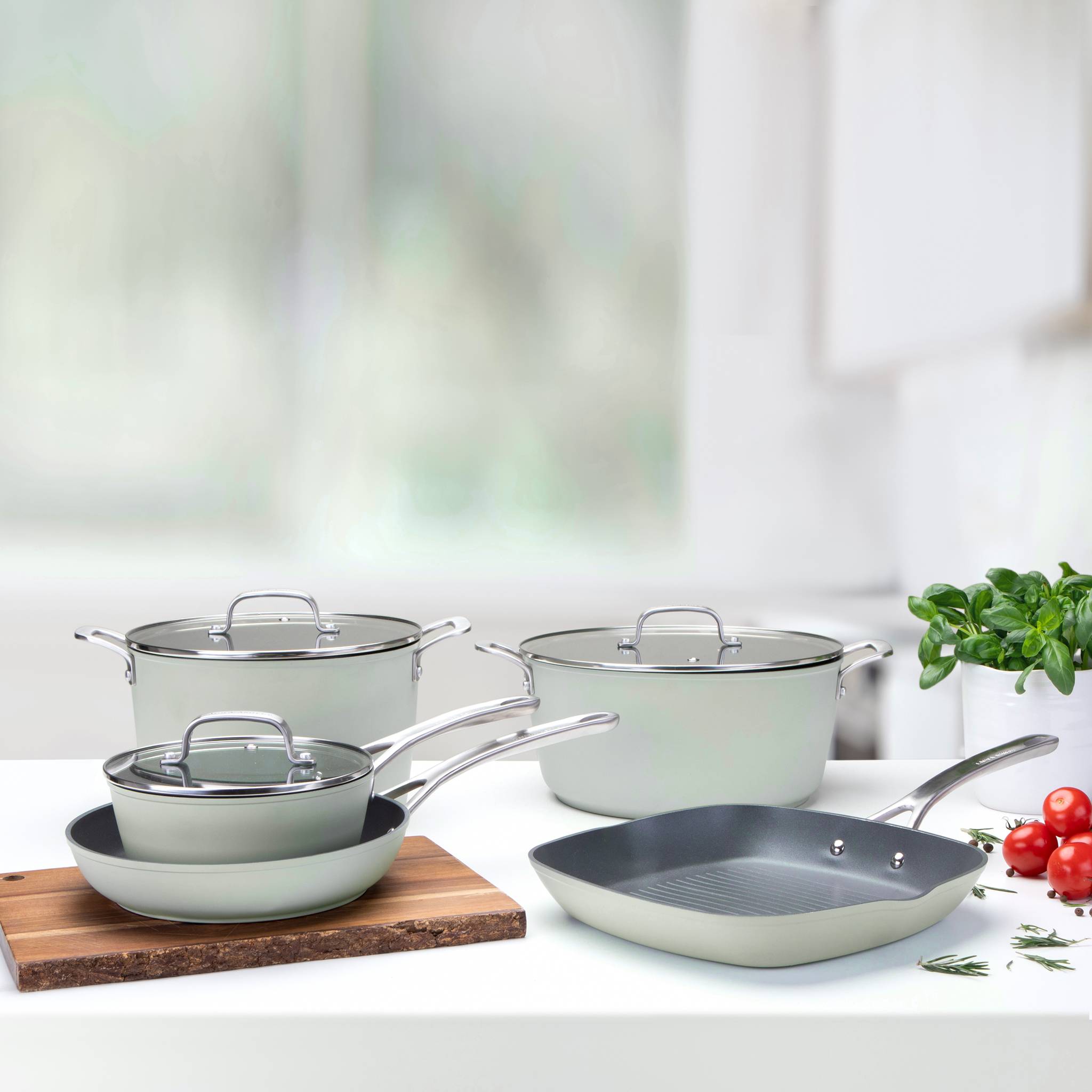 Ein Set aus grauer Kochware auf einer Küchenarbeitsplatte, bestehend aus Töpfen und Pfannen mit Glashdeckeln, neben Basilikumblättern.