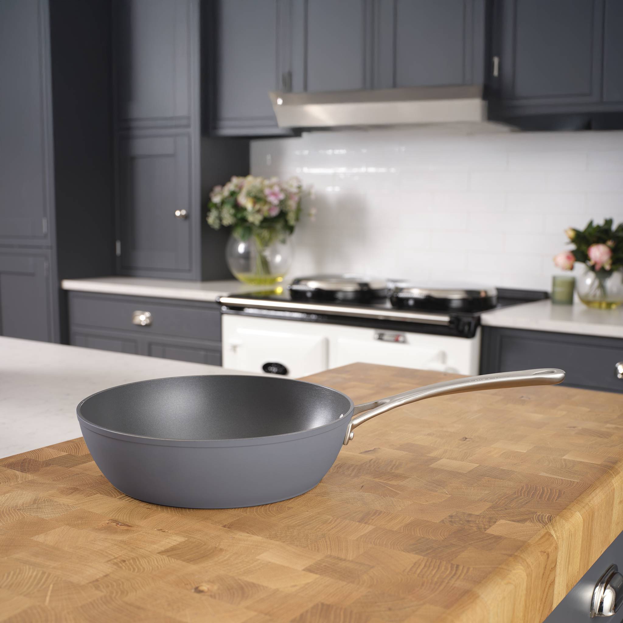 Eine antihaftbeschichtete Bratpfanne ruht auf einer hölzernen Kücheninsel. Der Küchenhintergrund zeigt weiße Schränke, einen Herd und Topfpflanzen.