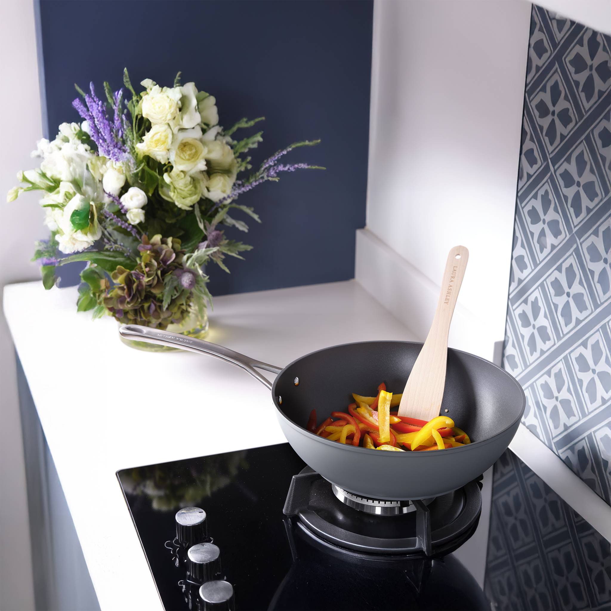 Eine Pfanne mit geschnittenen Paprikastücken steht auf einem Herd in einer modernen Küche. Ein Blumenstrauß sitzt in der Nähe und verleiht einen dekorativen Touch.