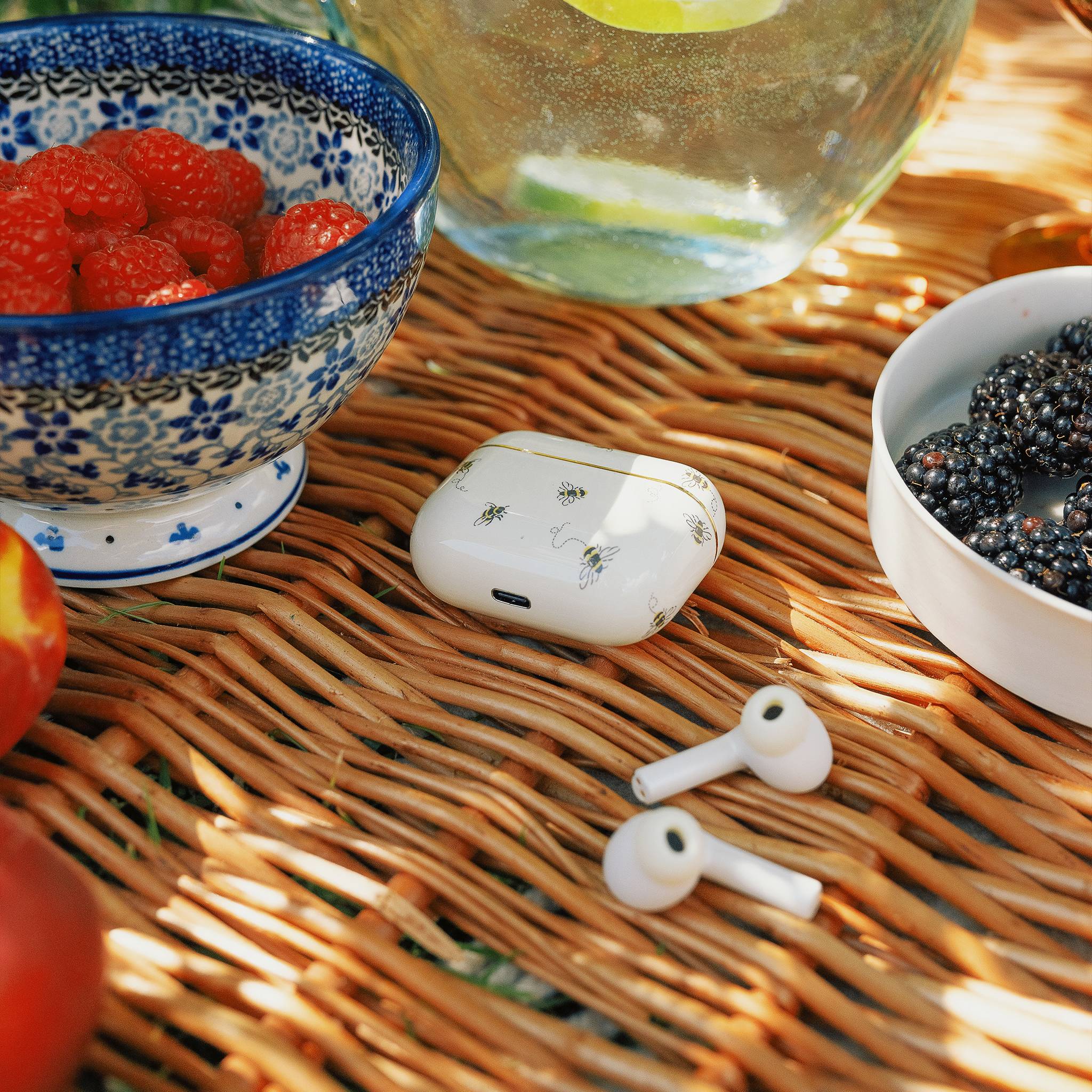 Eine Picknickszene auf einem Korbgeflecht-Tisch mit Beeren in Schüsseln, einem offenen Behälter für kabellose Ohrhörer, verziert mit kleinen Blumenillustrationen, und verstreuten Ohrhörern.