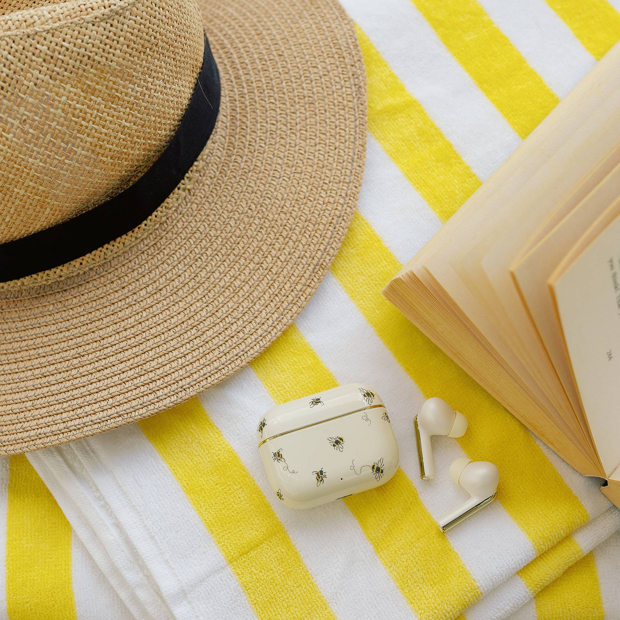 Ein Strohhut, ein aufgeschlagenes Buch und kabellose Kopfhörer in einem Ladeetui sind auf einem gelb-weiß gestreiften Stoff angeordnet und deuten auf eine entspannte Leseanlage hin.