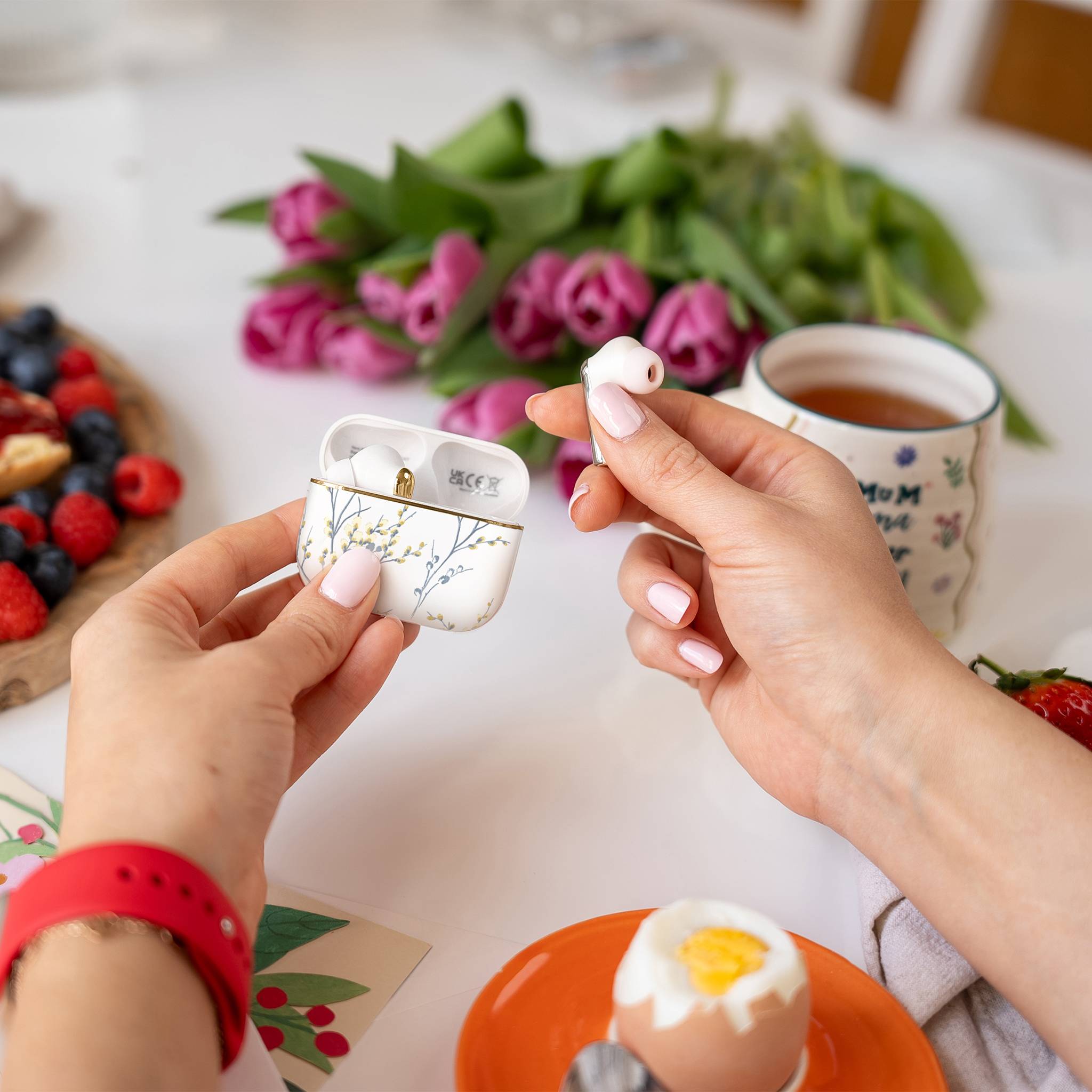 Eine Person, die einen kabellosen Ohrhörer in ein blumiges Etui auf einem Tisch mit einem angebissenen Ei, einer Tasse Tee, Beeren und rosa Tulpen in der Nähe legt.