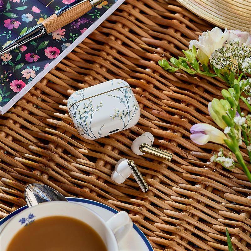 Kabellose Ohrhörer mit einem Gehäuse im Blumenmuster auf einer Korboberfläche, umgeben von einem Stift, einem Notizbuch mit Blumenmuster, Blumen und einer Kaffeetasse.