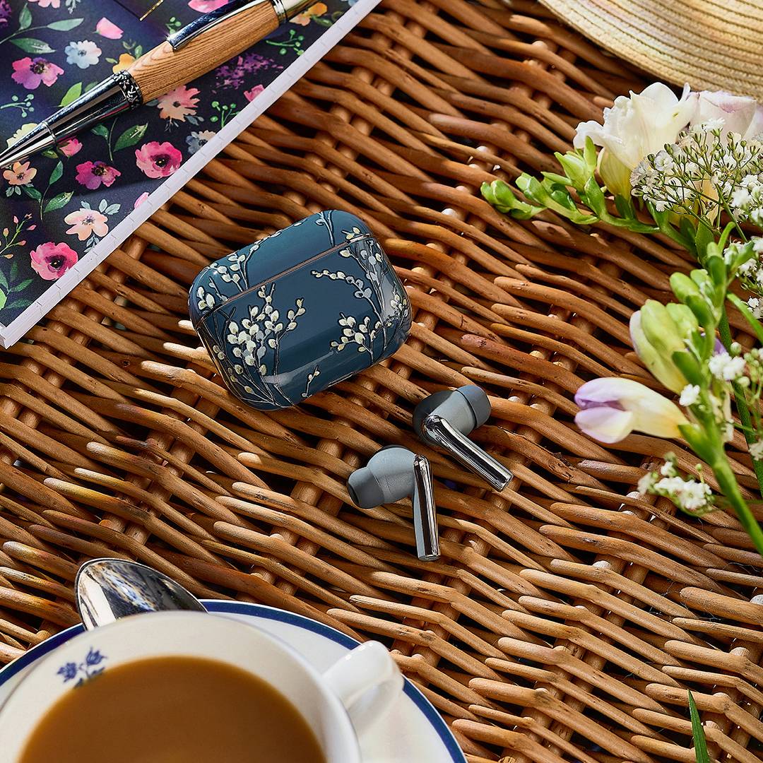 Kabellose Ohrhörer und ein Etui mit Blumenmuster auf einer Flechtoberfläche, umgeben von einem Notizbuch, einem Stift, einem Hut und einer Tasse Kaffee, mit Blumen in der Nähe.