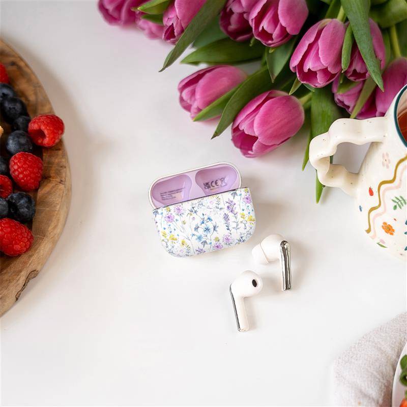 Kabellose Kopfhörer mit einem Gehäuse im Blumenmuster auf einer weißen Oberfläche, umgeben von einer Keramikkanne, frischen Tulpen und einem Teller mit Beeren.