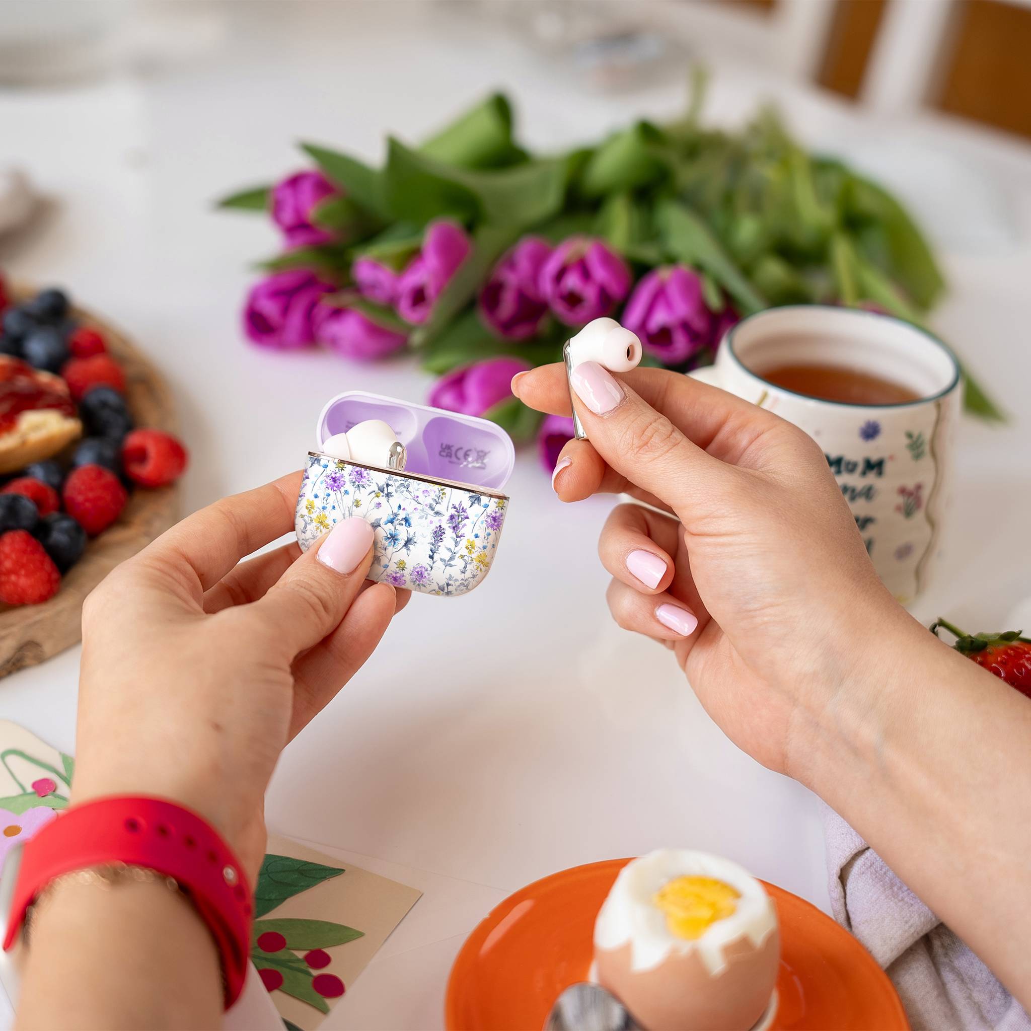 Eine Person hält ein kabelloses Ohrhörer in der Nähe einer offenen, mit Blumenmuster versehenen Ladeschale. Im Hintergrund befinden sich eine Teetasse, violette Blumen und ein Frühstückstablett.