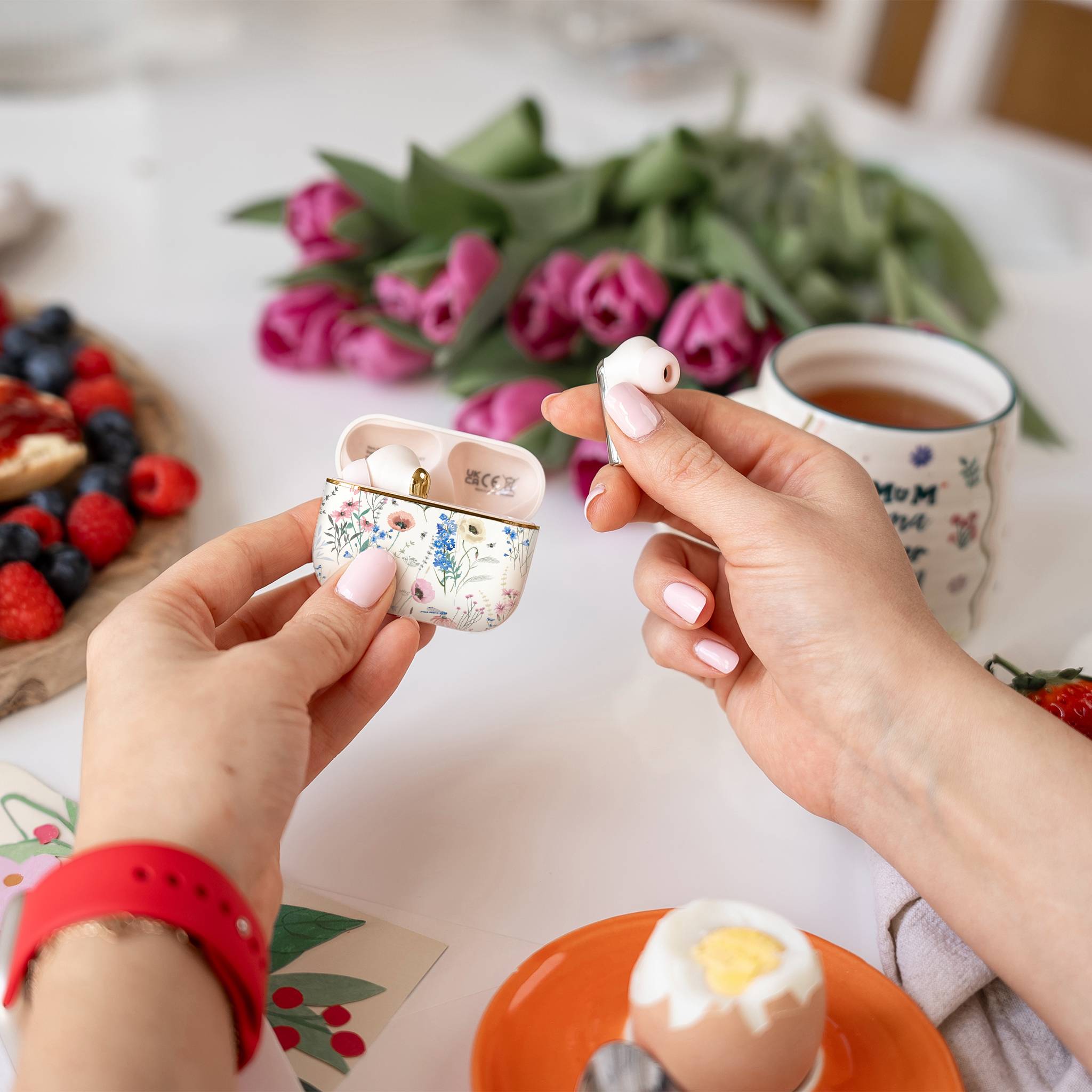 Eine Person hält blumenmusterige kabellose Ohrhörer über einen Frühstückstisch mit einem gekochten Ei, Beeren und einer Tasse Tee in der Nähe von rosa Tulpen.
