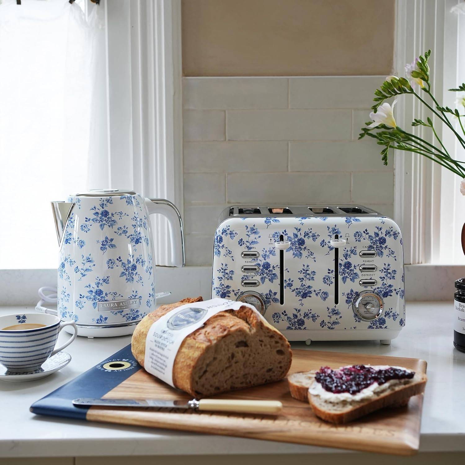 Eine Küchenarbeitsplatte mit einem blumigen Toaster und einem passenden Wasserkocher neben geschnittenem Brot, einer Marmeladenbrotschnitte mit Butter und einer Tasse mit Untertasse.