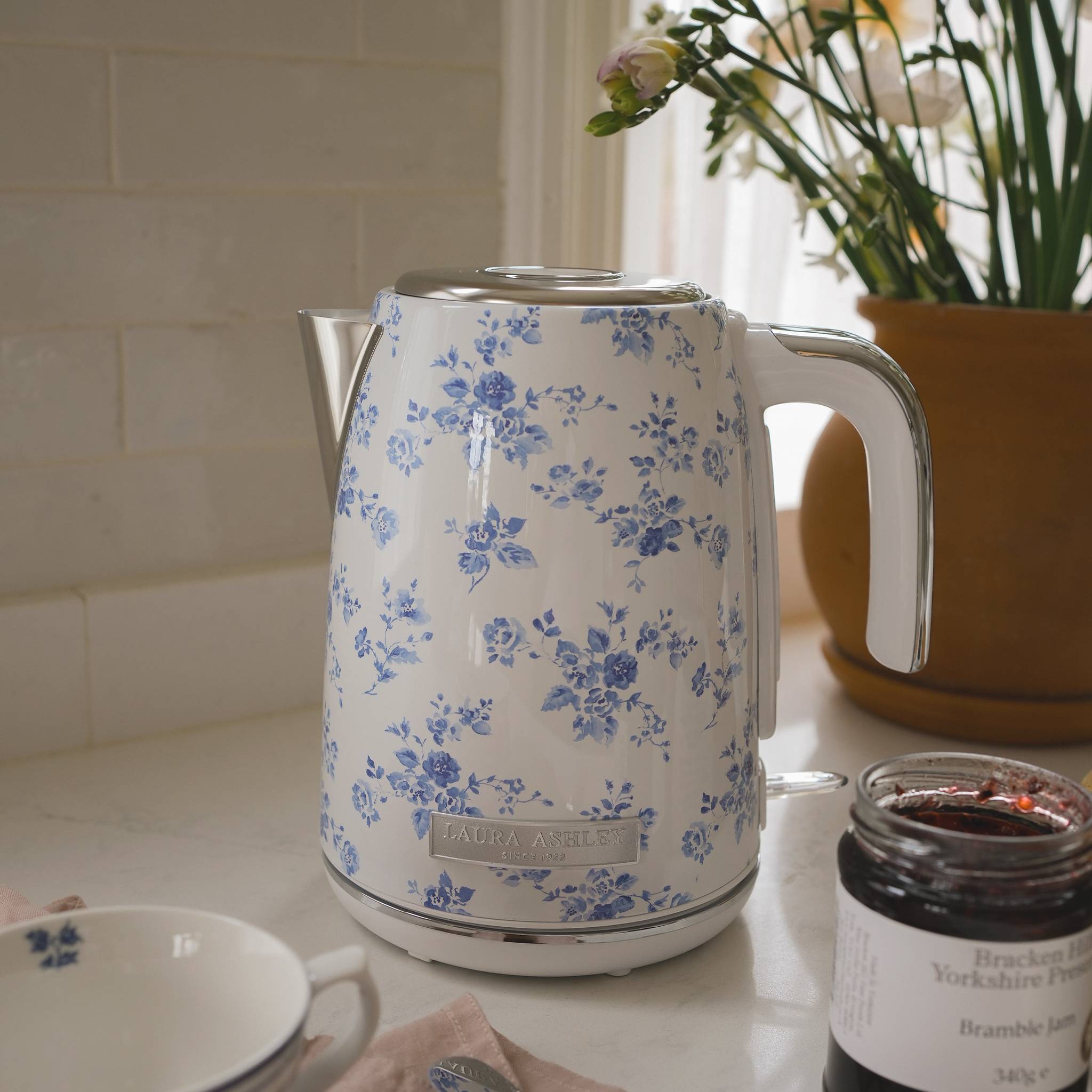 Ein elektrischer Wasserkessel mit Blumenmuster steht auf einer Küchenarbeitsplatte, umgeben von einer Topfpflanze, einem Teller, einem Löffel und einem offenen Marmeladenglas.