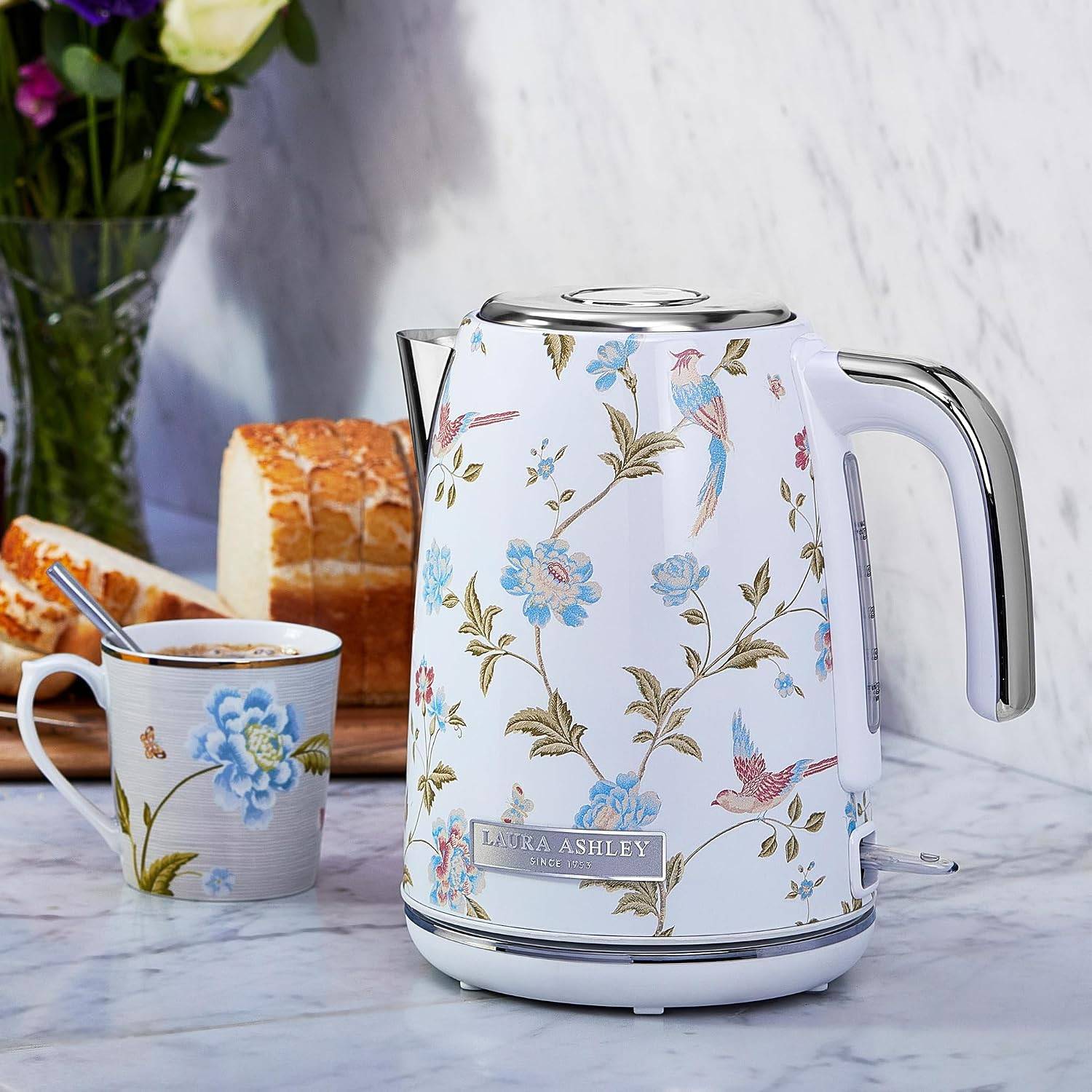 Elektrischer Wasserkessel mit Blumenmuster und passende Tasse auf einer Marmorarbeitsfläche neben geschnittenem Brot und Blumen.