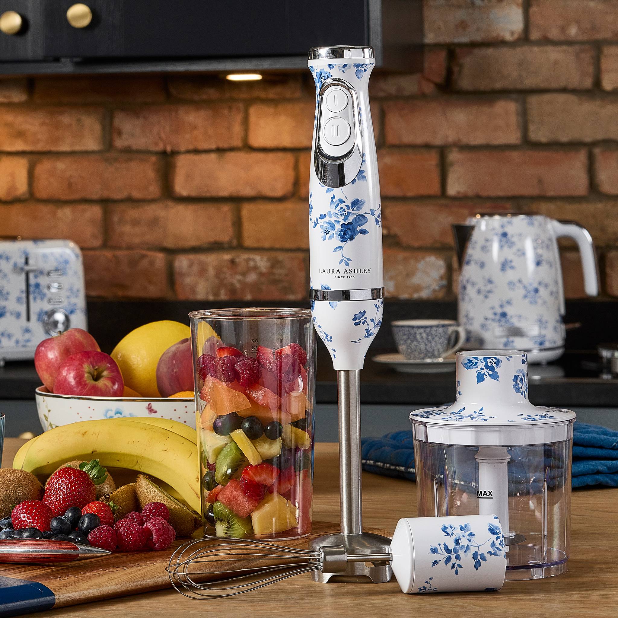 Ein handmixer mit floralem Muster steht auf einer Küchentheke, umgeben von frischem Obst, einem fruchtgefüllten Glas und Küchenutensilien.