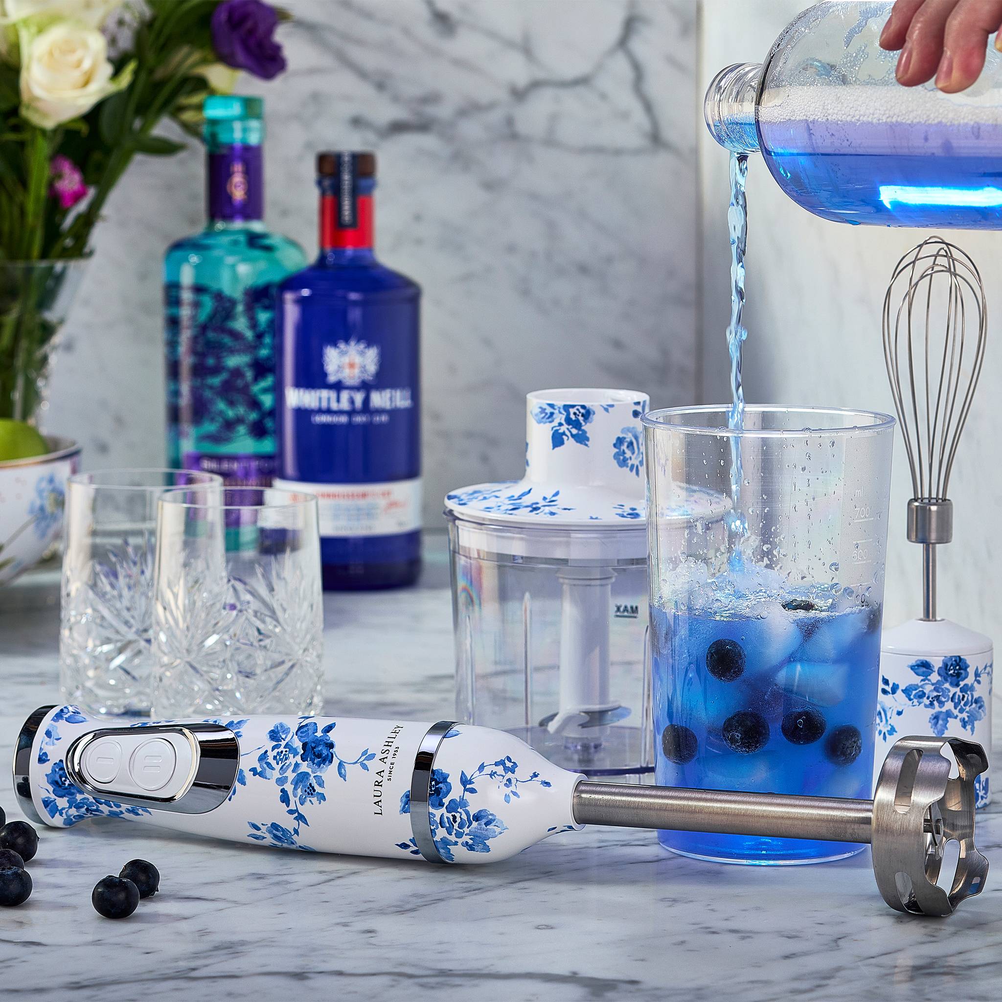Eine Person gießt blaue Flüssigkeit aus einem Streuer in ein Glas mit Blaubeeren auf einer Marmortheke, umgeben von blauen Blumenküchengeräten.